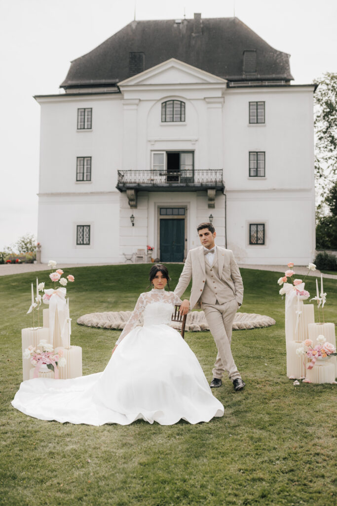 Die Braut in einem weißen Kleid und der Bräutigam in einem beigen Anzug posieren vor einem großen weißen Gebäude mit Blumendekor, das die Eleganz einer Hochzeit in Süddeutschland einfängt.