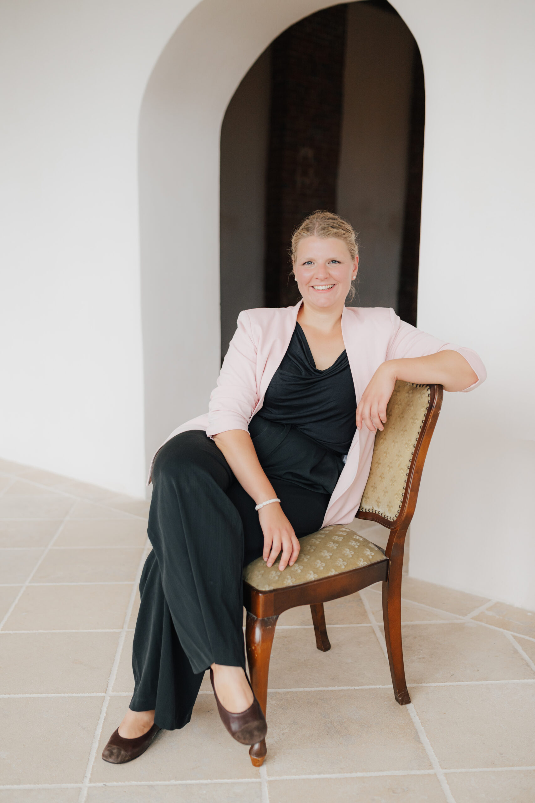 Stephanie Menzdorf in einem rosa Blazer und schwarzem Outfit sitzt auf einem Vintage-Stuhl in einem hellen, gewölbten Raum - die perfekte Inspiration für jede Hochzeitsplanerin in Süddeutschland oder Österreich.