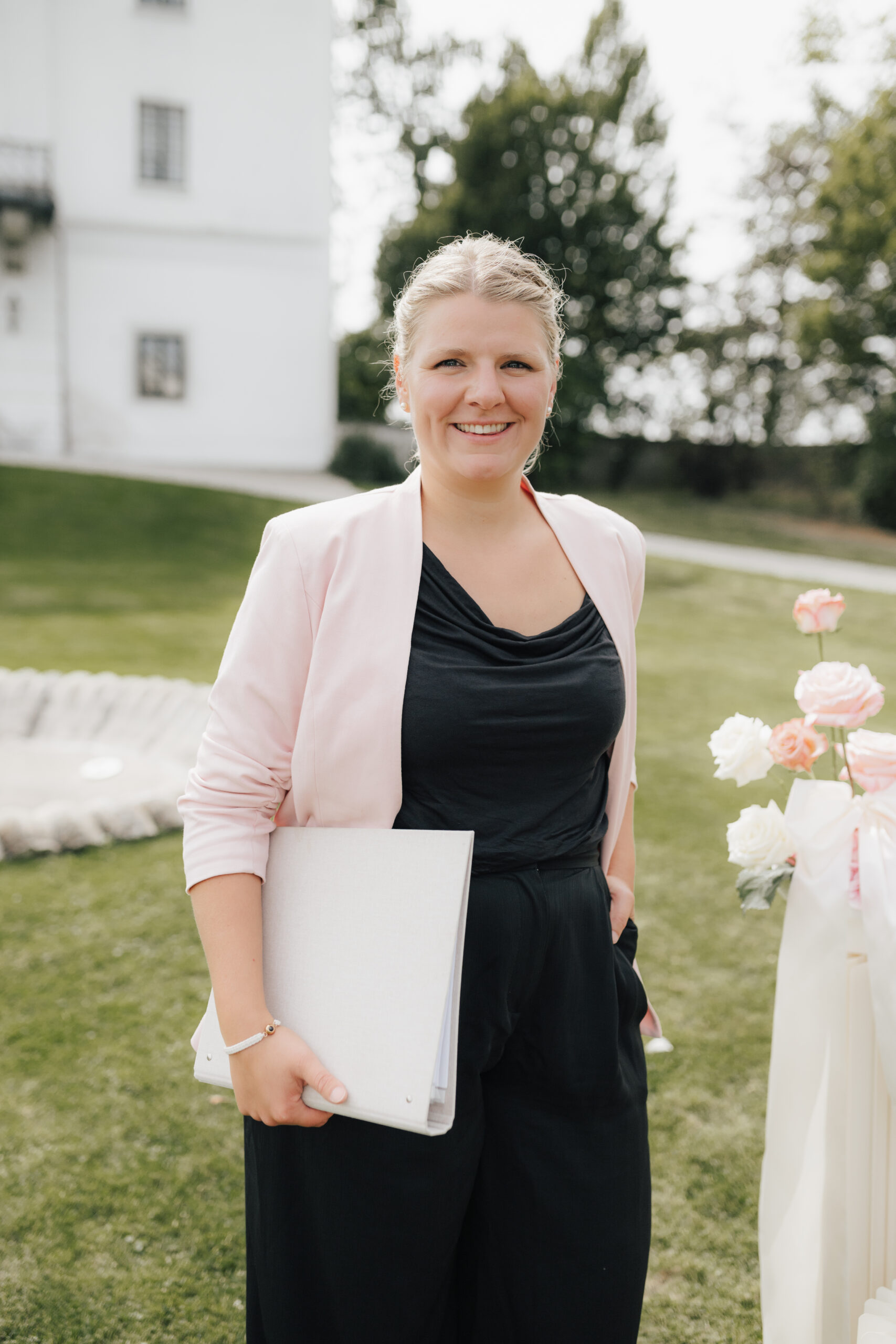 Lachende vrouw, Stephanie Menzdorf, in een roze blazer, met een map in haar hand, staat buiten op een zonnige dag bij bloemen, mogelijk op een bruiloft in Zuid-Duitsland of Oostenrijk.