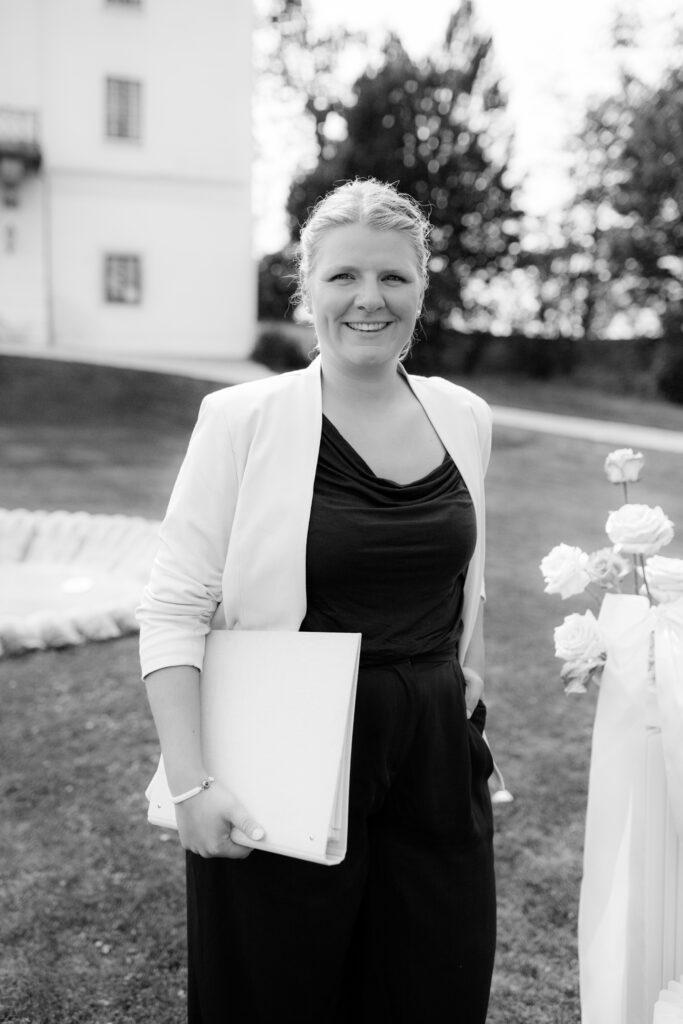 Stephanie Menzdorf, wedding planner in a blazer with a folder in her hand, standing outdoors next to flowers and a building in Austria.