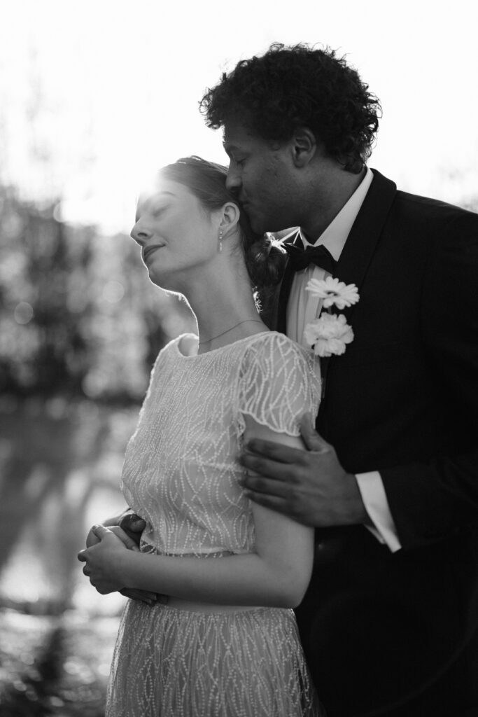 A groom gently kisses his bride's head outdoors in Southern Germany, both in formal wedding attire, with soft sunlight in the background – beautifully captured by an experienced wedding planner.