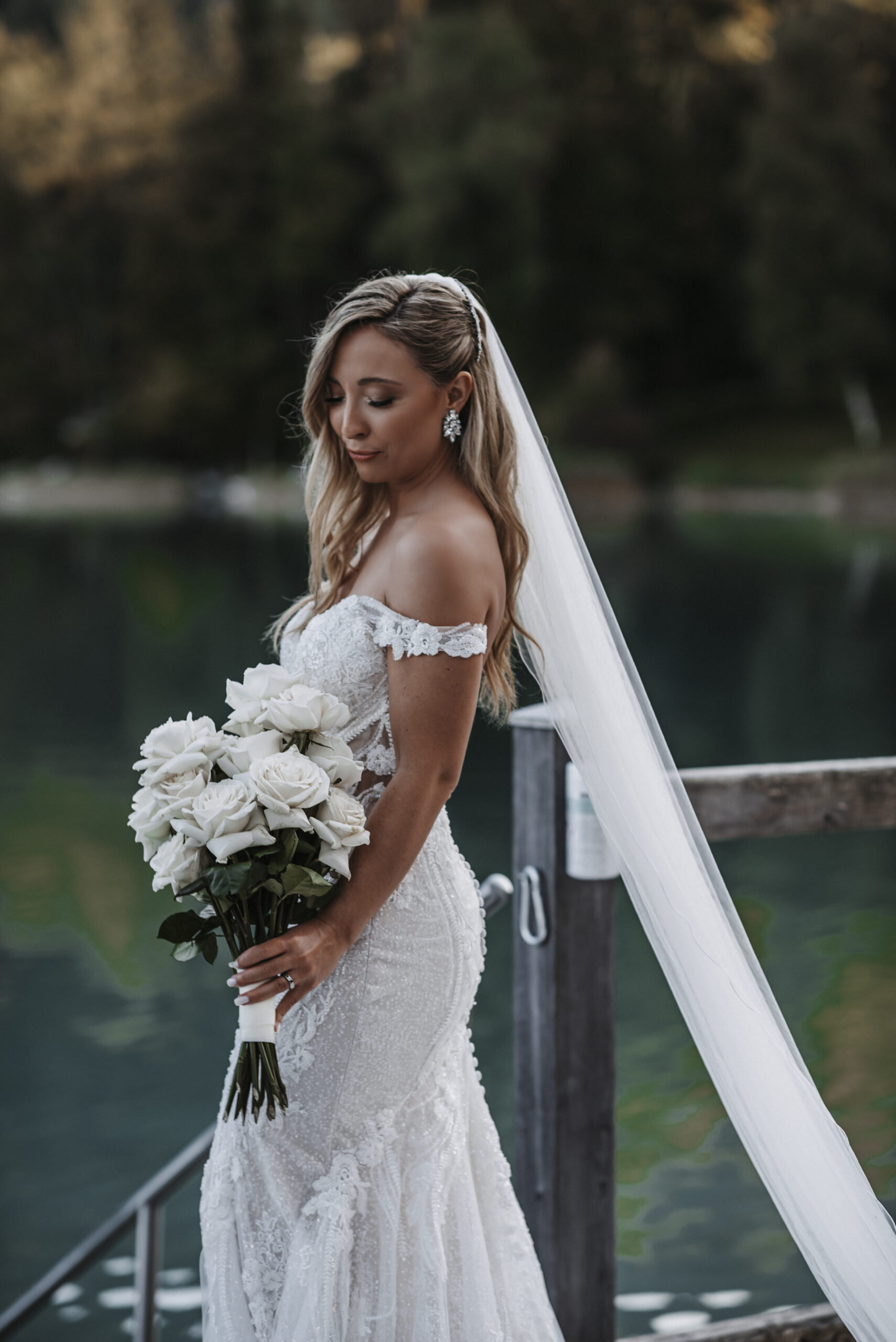The bride in a white lace dress with white roses in her hand stands by a wooden railing near a lake – the perfect inspiration for your wedding in Southern Germany, carefully curated by an experienced wedding planner.