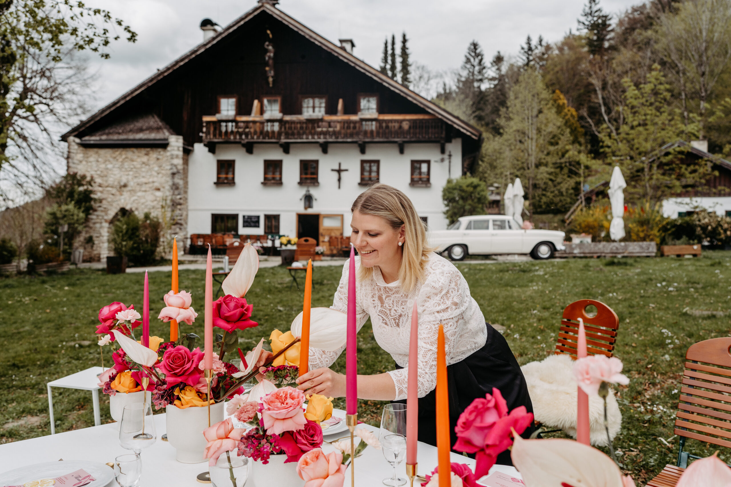 Eine Hochzeitsplanerin arrangiert leuchtende Blumen auf einem Tisch im Freien vor einem rustikalen Haus mit Garten und fängt damit den Charme Süddeutschlands ein.
