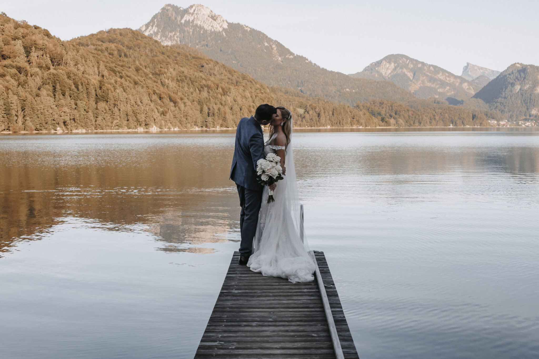 Braut und Bräutigam küssen sich auf einem hölzernen Steg an einem ruhigen See mit Bergen im Hintergrund - eine bezaubernde Szene, perfekt für jede Hochzeitsplanerin in Österreich oder Süddeutschland.