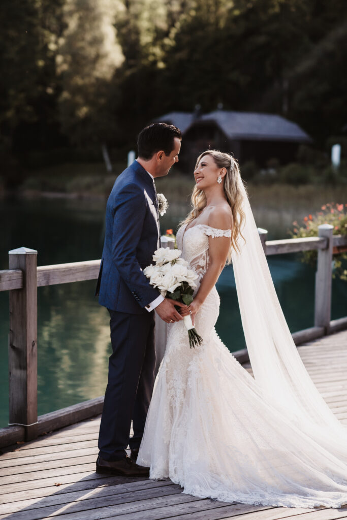 Braut und Bräutigam lächeln sich auf einem hölzernen Steg am Wasser an, die Braut hält einen Blumenstrauß in der Hand - so wird der Zauber einer Hochzeit in Süddeutschland eingefangen.