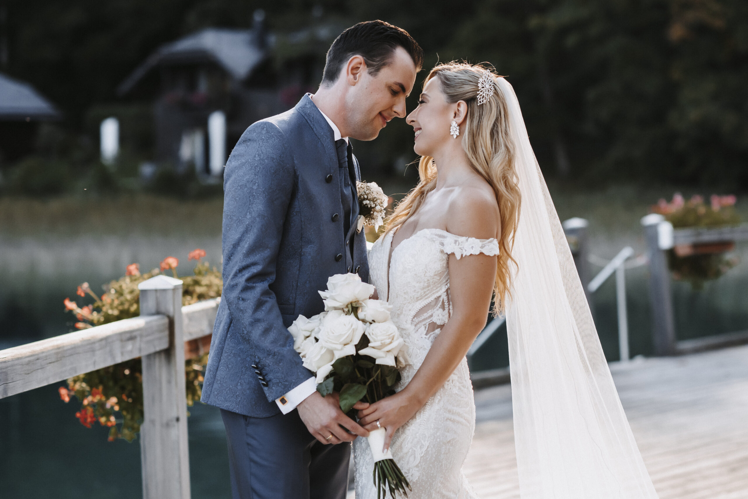 Braut und Bräutigam lächeln sich auf einem hölzernen Steg in Süddeutschland an, halten weiße Rosen in der Hand und haben Bäume im Hintergrund - perfekt für eine romantische Hochzeit.