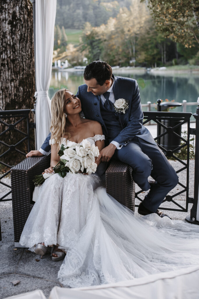 Eine Braut und ein Bräutigam sitzen im Freien an einem See in Süddeutschland und lächeln sich liebevoll an, um den Zauber ihrer Hochzeit einzufangen.