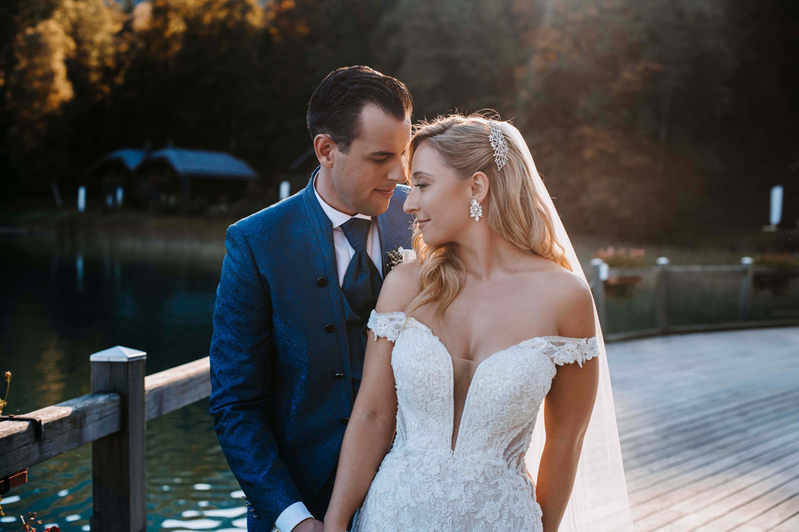 Braut und Bräutigam in eleganter Hochzeitskleidung umarmen sich im Freien an einem See in Süddeutschland, getaucht in sanftes Sonnenlicht - ein unvergesslicher Hochzeitsmoment.