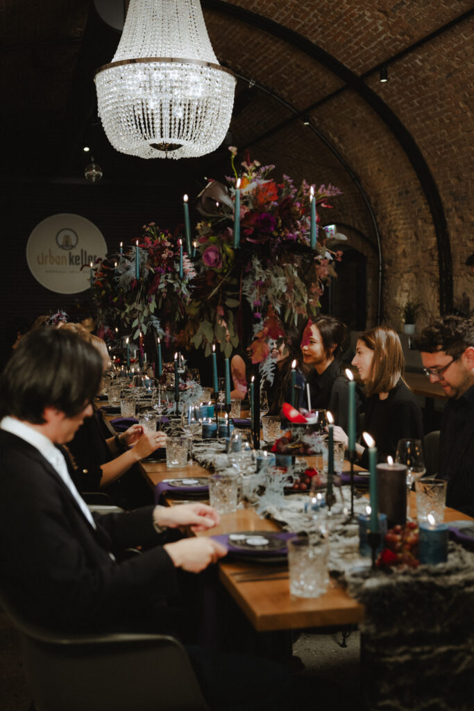 Menschen an einem langen, elegant gedeckten Tisch mit Kerzen und Blumen unter einem großen Kronleuchter in einem gemauerten Raum, perfekt für eine unvergessliche Hochzeitsplanerin Veranstaltung in Österreich oder Süddeutschland.