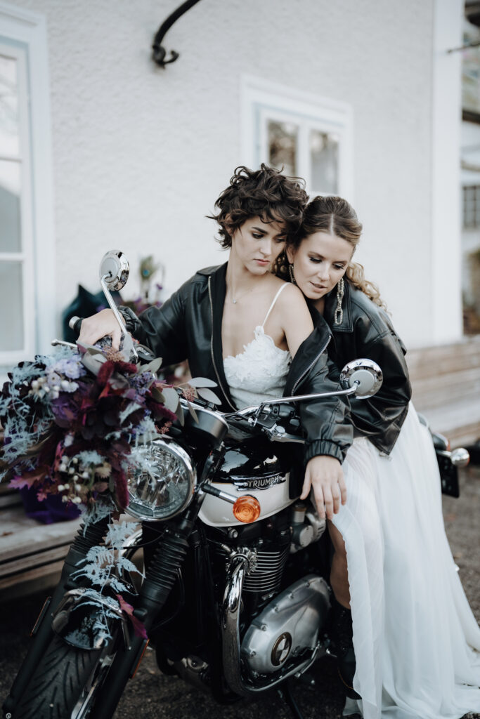 Zwei Bräute in weißen Kleidern und schwarzen Jacken sitzen auf einem mit lila Blumen geschmückten Motorrad und feiern ihre Liebe mit Stil - die perfekte Inspiration für eine Hochzeitsplanerin in Österreich oder Süddeutschland.