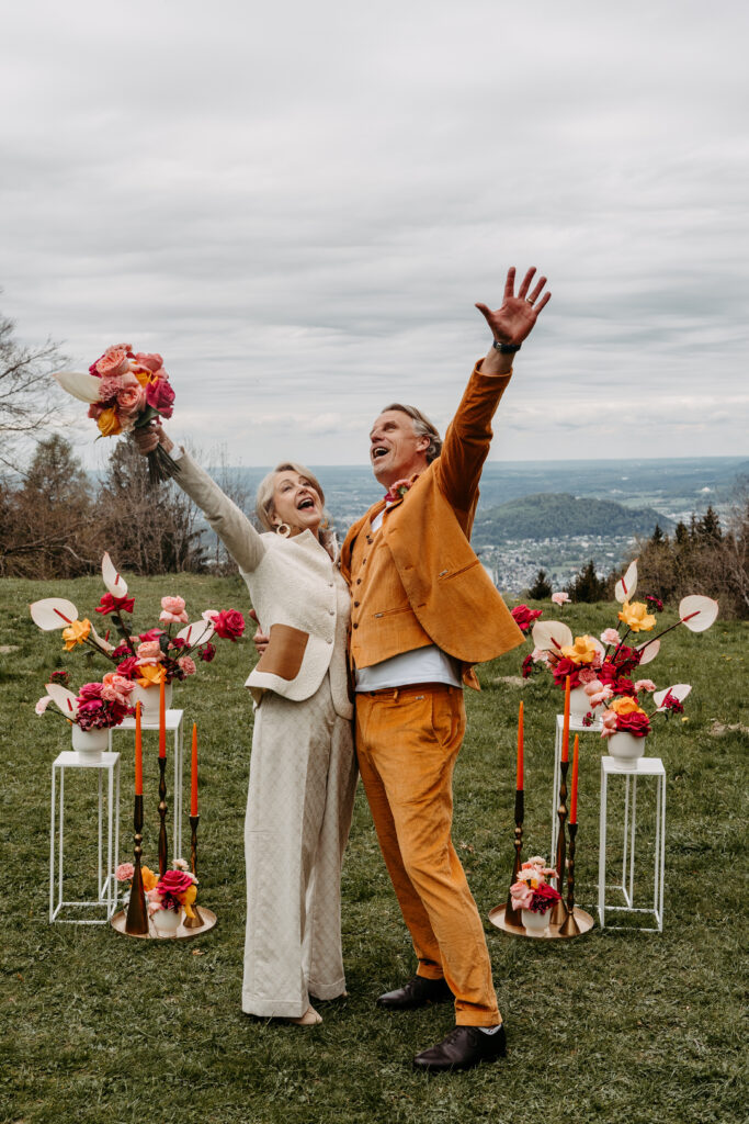 Zwei fröhliche Menschen in Anzügen feiern ihre Hochzeit im Freien in Süddeutschland, umgeben von bunten Blumen und eleganten Kerzenarrangements.