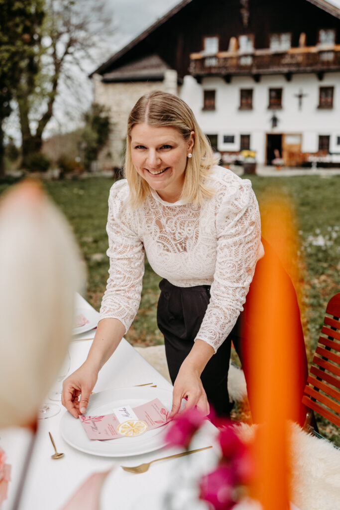 A woman in a white lace top smiles as she sets an outdoor table, with a house in the background - perfect for a wedding in Southern Germany.