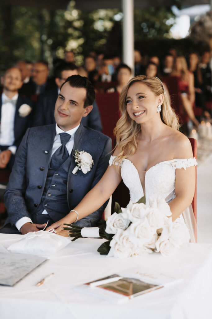 Braut und Bräutigam sitzen während ihrer Hochzeit lächelnd an einem Tisch, halten sich an den Händen und sind von Gästen umgeben, während ihre Hochzeitsplanerin dafür sorgt, dass jedes Detail perfekt ist.