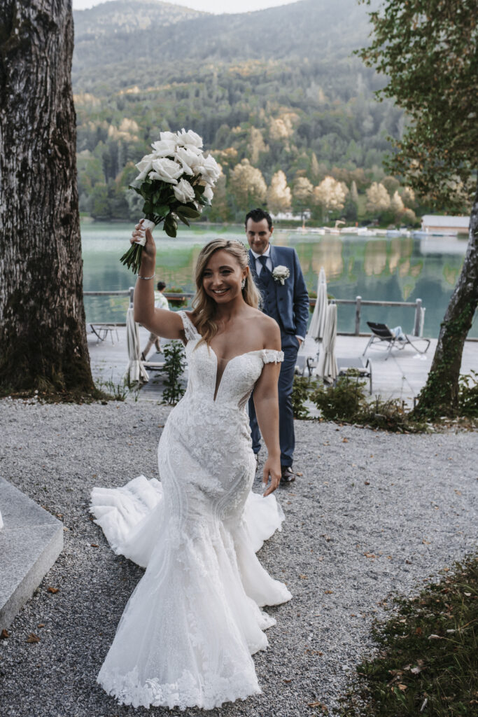 Eine lächelnde Braut in einem weißen Kleid hält ihren Brautstrauß in die Höhe, der Bräutigam im Anzug steht hinter ihr an einem See - ein wunderschöner Moment, der oft von einer erfahrenen Hochzeitsplanerin aus Österreich oder Süddeutschland gestaltet wird.