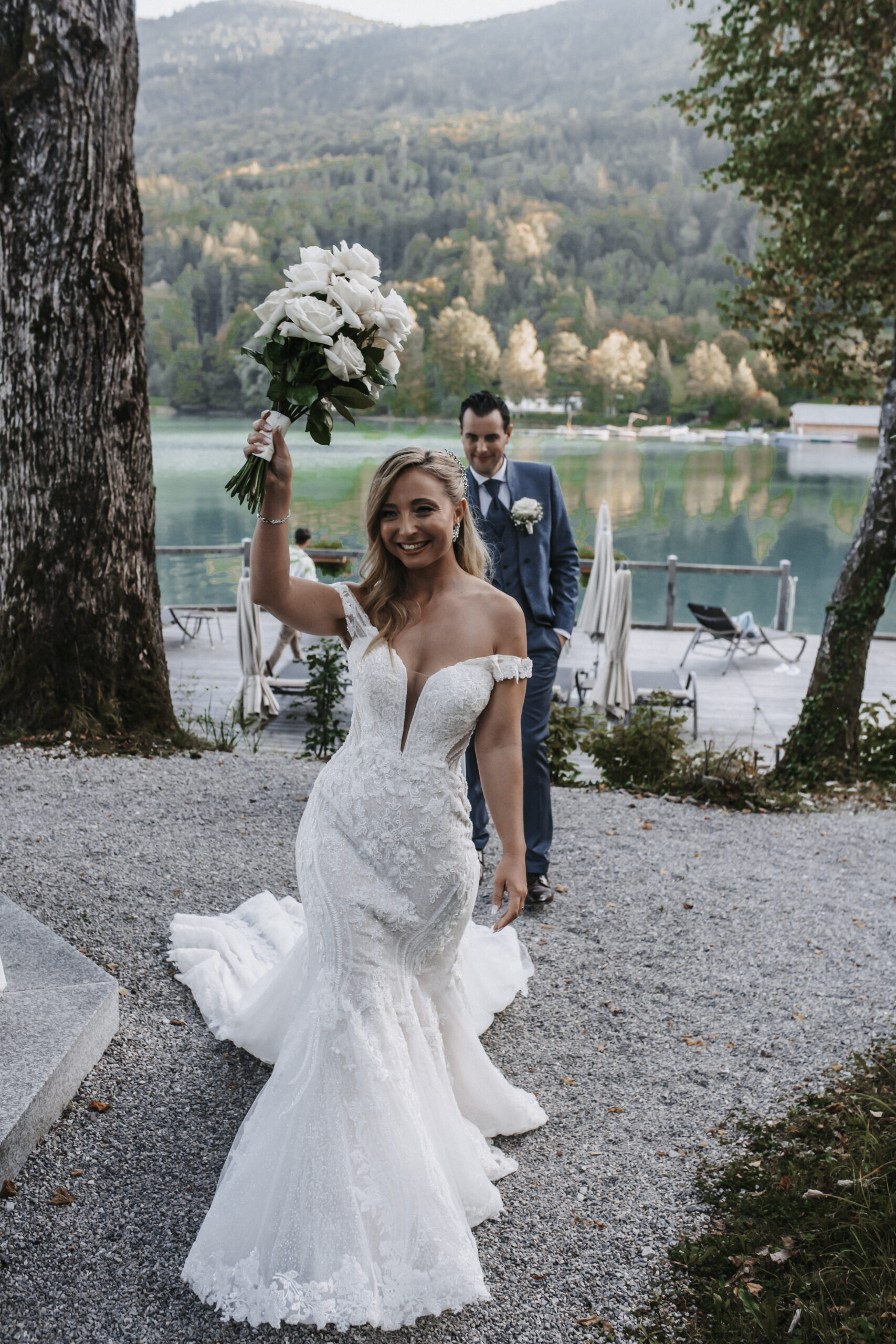 Eine lächelnde Braut in einem weißen Kleid hält ihren Brautstrauß in die Höhe, der Bräutigam im Anzug steht hinter ihr an einem See - ein wunderschöner Moment, der oft von einer erfahrenen Hochzeitsplanerin aus Österreich oder Süddeutschland gestaltet wird.
