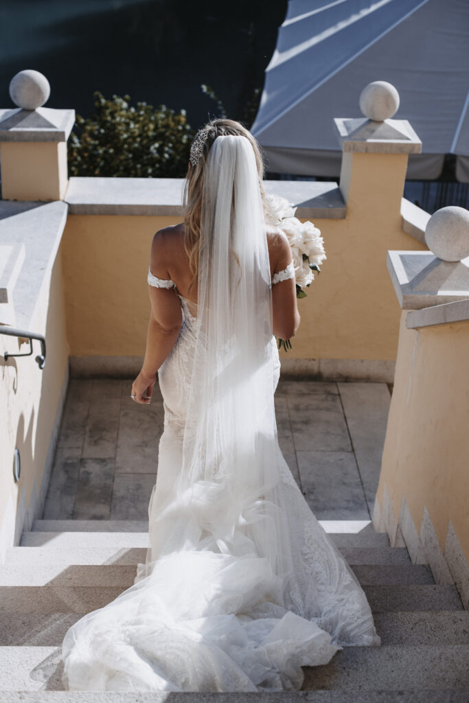 Die Braut in weißem Kleid und Schleier, mit Blumen in der Hand, schreitet die sonnenbeschienenen Steinstufen im Freien hinauf - ein eleganter Moment, perfekt für eine Hochzeitsplanerin in Österreich oder Süddeutschland.