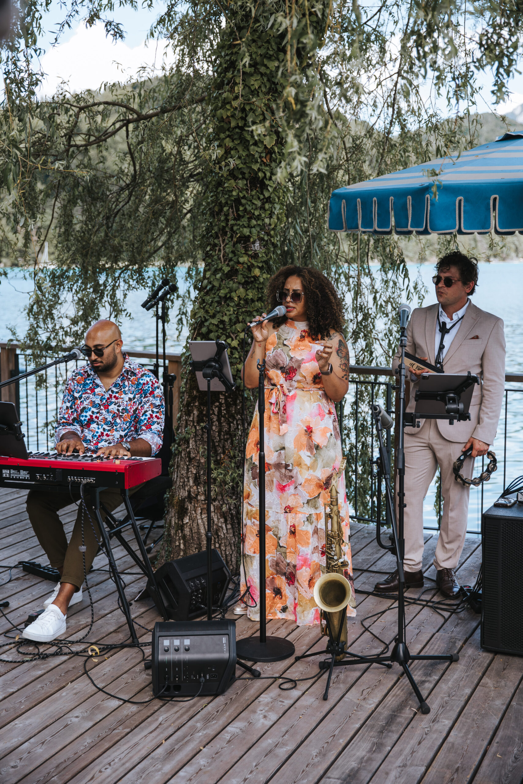 Three musicians are playing outdoors by a lake in Southern Germany; one is playing a keyboard, one is singing, and one is holding a saxophone.