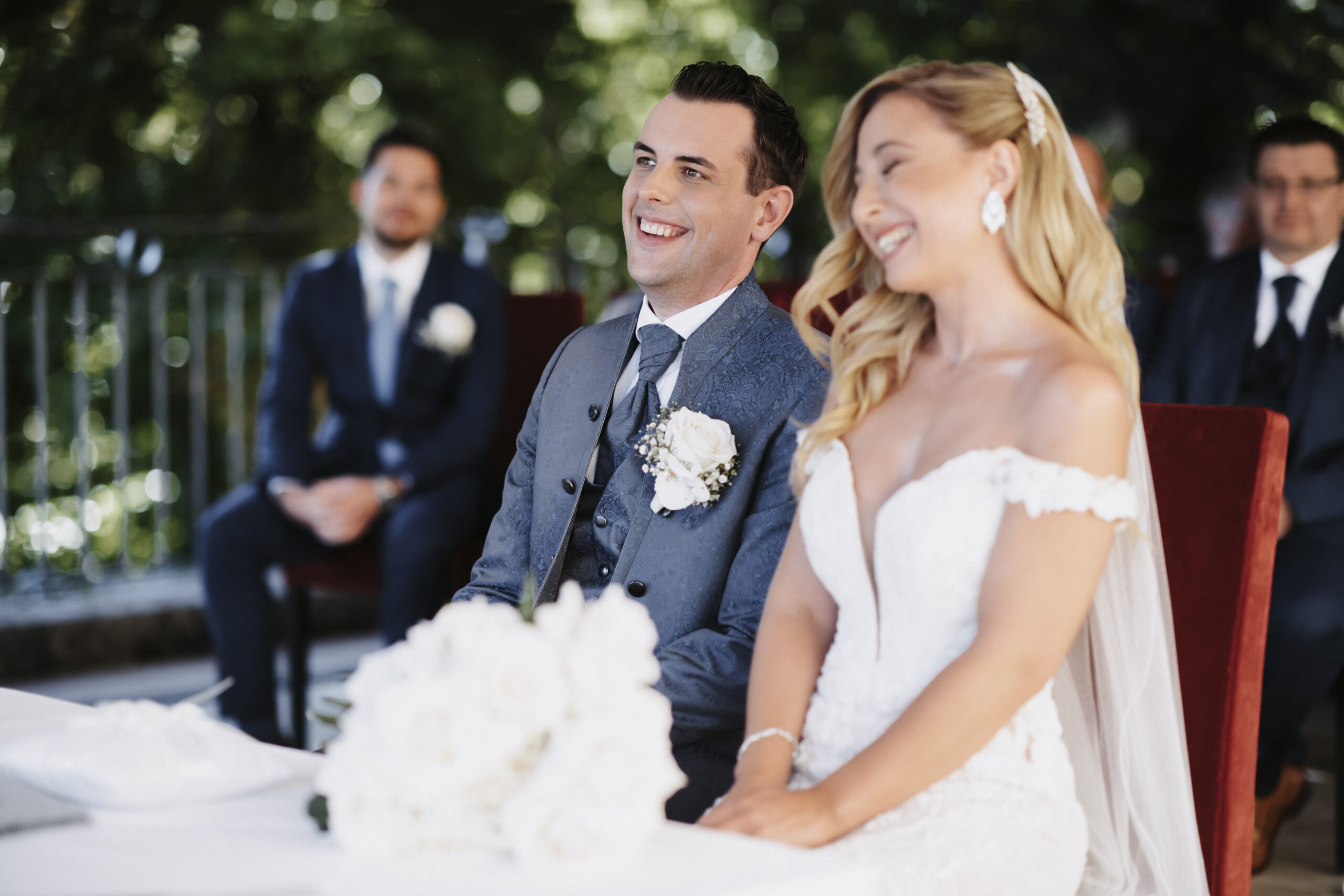 Lächelnde Braut und Bräutigam sitzen bei ihrer Hochzeit in Österreich Seite an Seite, dahinter sitzen Gäste und ihre Hochzeitsplanerin.