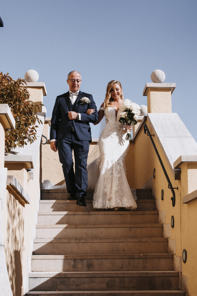 Die Braut in einem Spitzenkleid geht am Arm ihres Vaters bei ihrer Hochzeit die Freitreppe hinunter, beide lächeln unter einem klaren blauen Himmel in Süddeutschland.