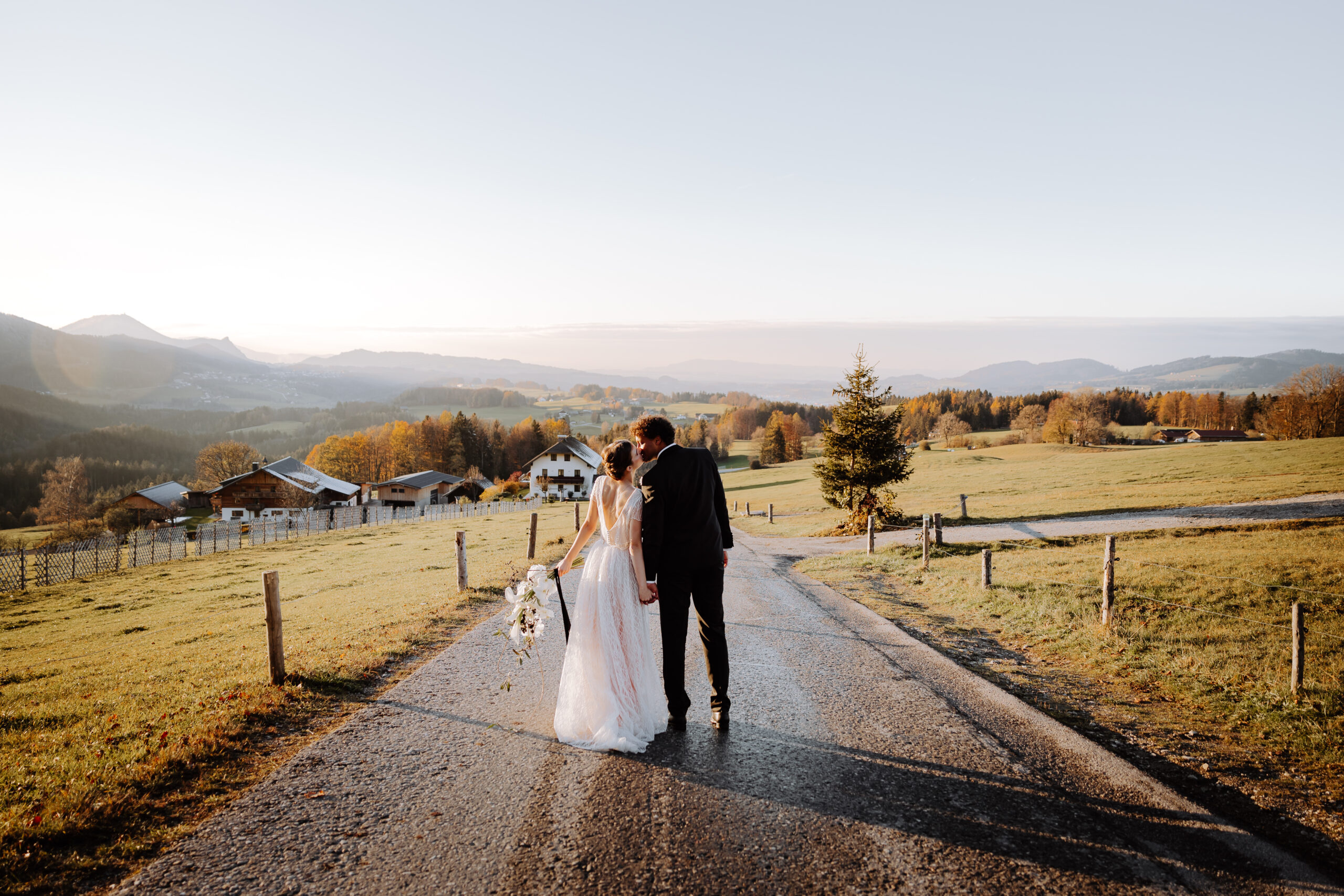 Eine Braut und ein Bräutigam gehen bei Sonnenuntergang eine Landstraße entlang, Heiraten in Österreich mit bergpanorama, Hochzeitslocation Ansitz Wartenfels nähe Salzburg