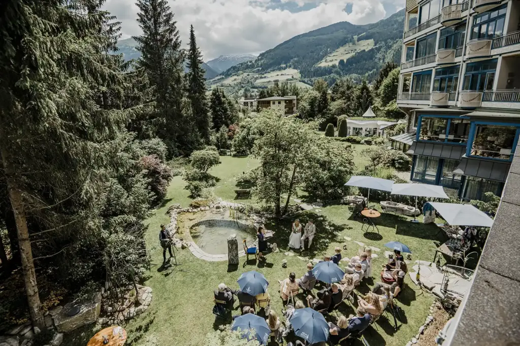 Hochzeitszeremonie im Freien in einem üppigen Garten, umgeben von Bergen - eine Traum-Hochzeitslocation in Österreich, wo die Gäste unter Sonnenschirmen inmitten einer atemberaubenden Kulisse Platz nehmen.