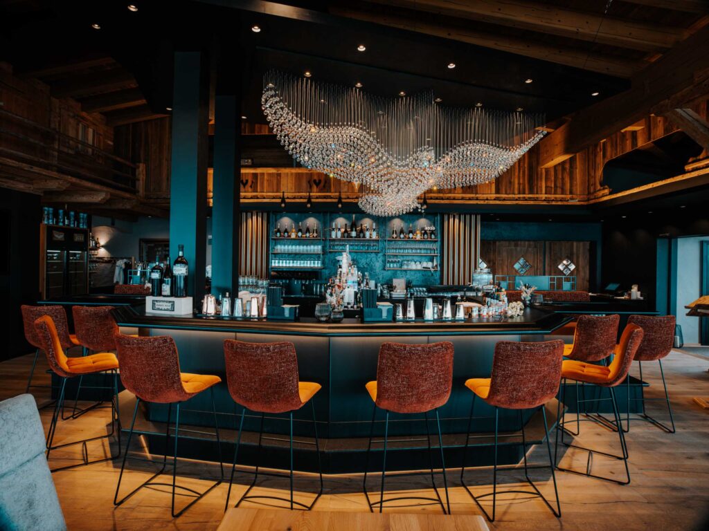 Eine moderne Bar mit orangefarbenen Stühlen, einem großen Tresen in der Mitte und einem auffälligen Kronleuchter in Form einer geflügelten Figur - eine unvergessliche Hochzeitslocation in Österreich für Paare, die eine stilvolle und einzigartige Feier suchen. Hochzeitslocation Jazz AlmResort in Tirol