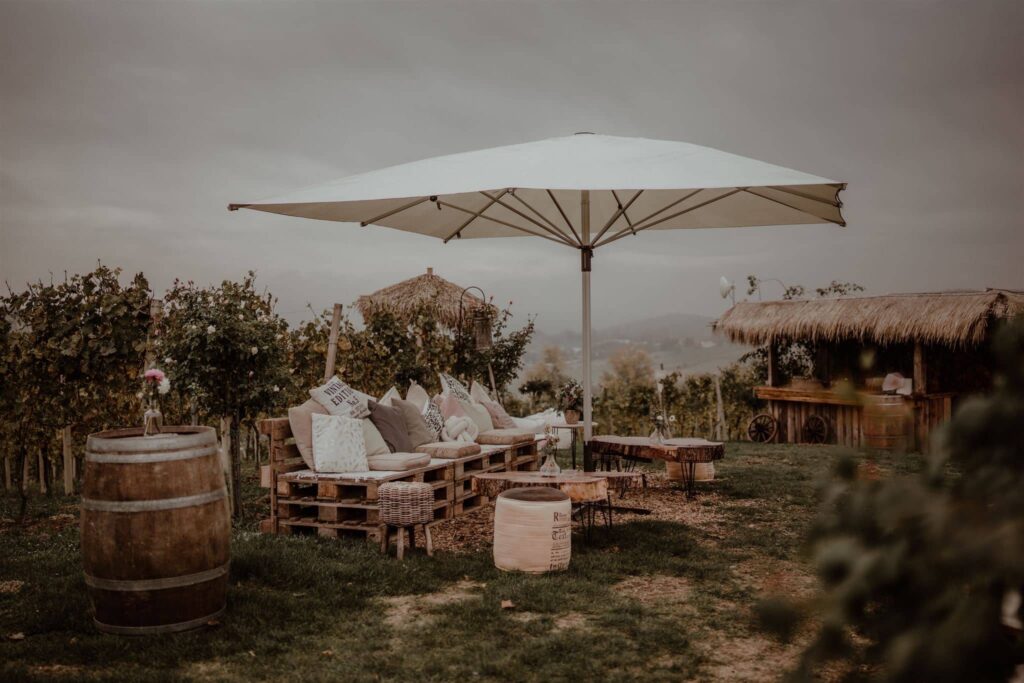 Outdoor-Lounge mit Palettensofas, Kissen, großem Sonnenschirm, Weinfässern und einer rustikalen Bar inmitten eines Weinberges, Hochzeitslocation Steiermark Weingut Holler