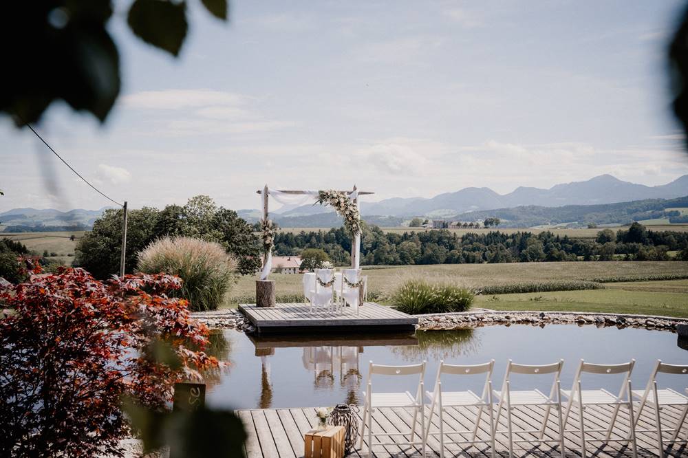 Ein Hochzeitsaltar im Freien an einem Teich mit weißen Stühlen, Bergblick und klarem Himmel - die perfekte Hochzeitslocation in Österreich für Paare, die von einer romantischen Zeremonie inmitten der Natur träumen.