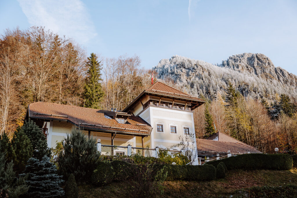 Charmantes weißes Haus mit rotem Dach, umgeben von Bäumen, vor einem felsigen Berg und blauem Himmel - eine idyllische Hochzeitslocation in Österreich für Brautpaare, die von Heiraten in Österreich träumen. Hochzeitslocation in Österreich Ansitz Wartenfels