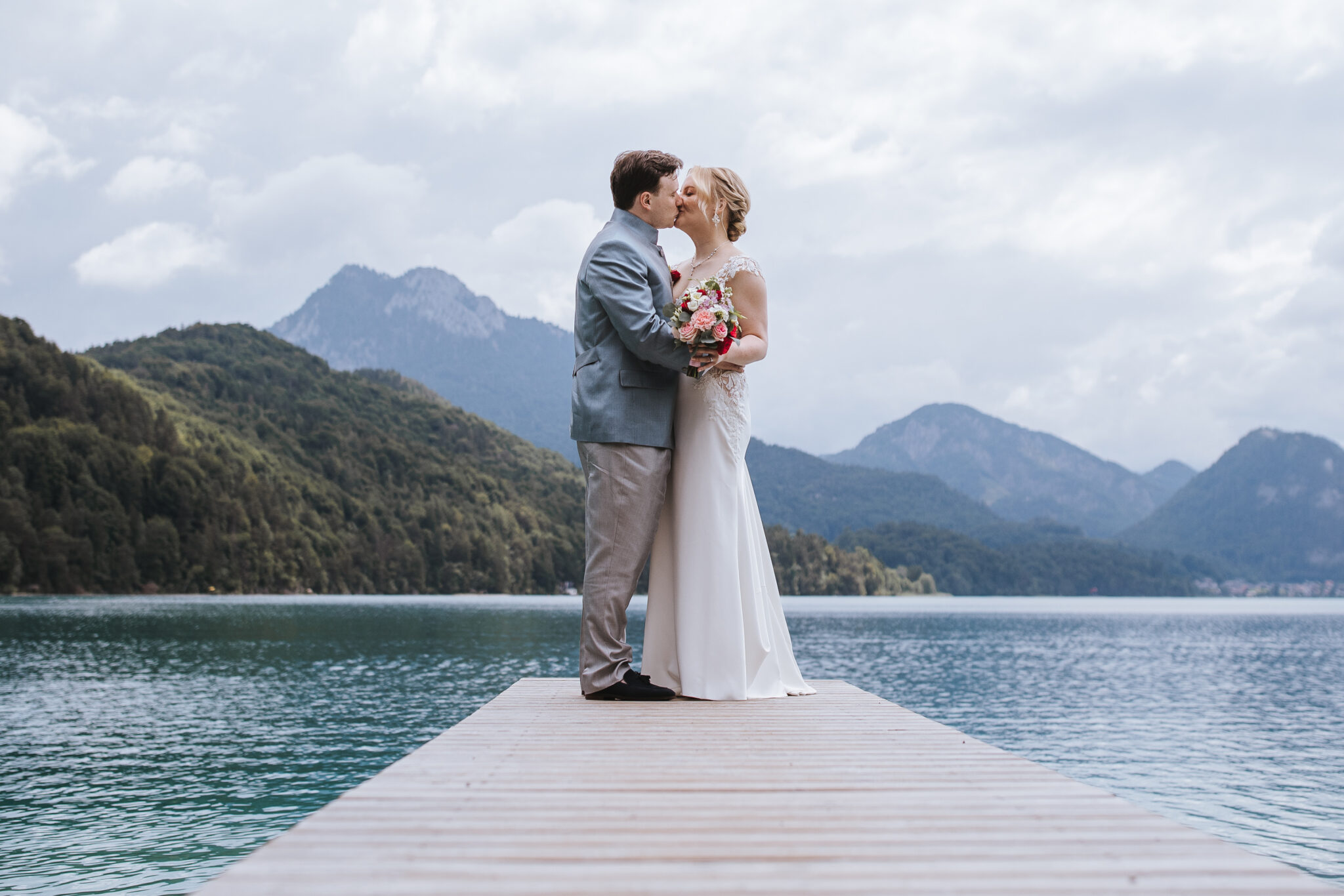 Braut und Bräutigam küssen sich auf einem Holzsteg vor dem Hintergrund von Bergen und einem See – eine romantische Hochzeit an einem besonderen Ort in Österreich.
