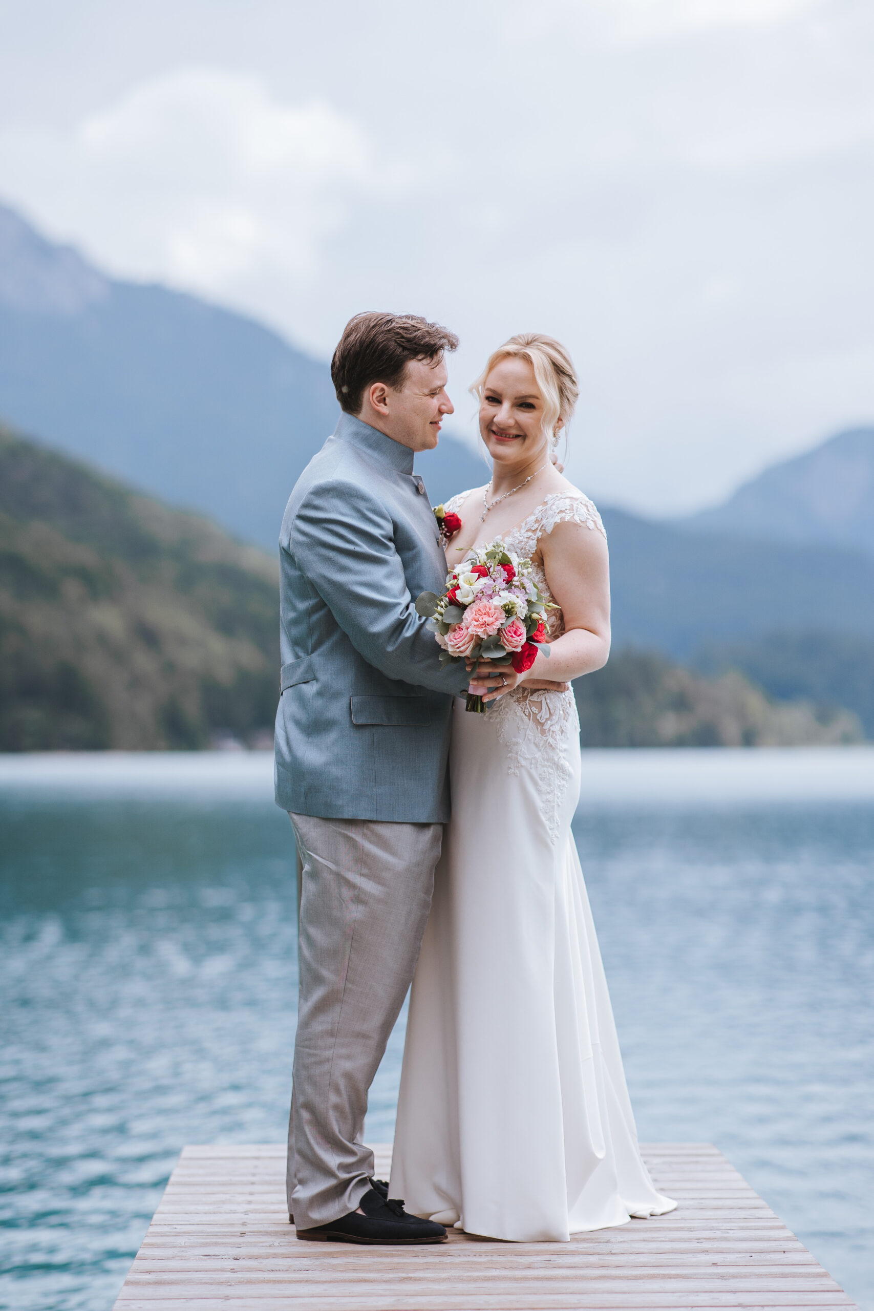 Braut und Bräutigam stehen auf einem Steg an einem See in Österreich, umarmen sich und lächeln, im Hintergrund sind Berge zu sehen – ein perfekter Moment für eine Hochzeit im Ausland.