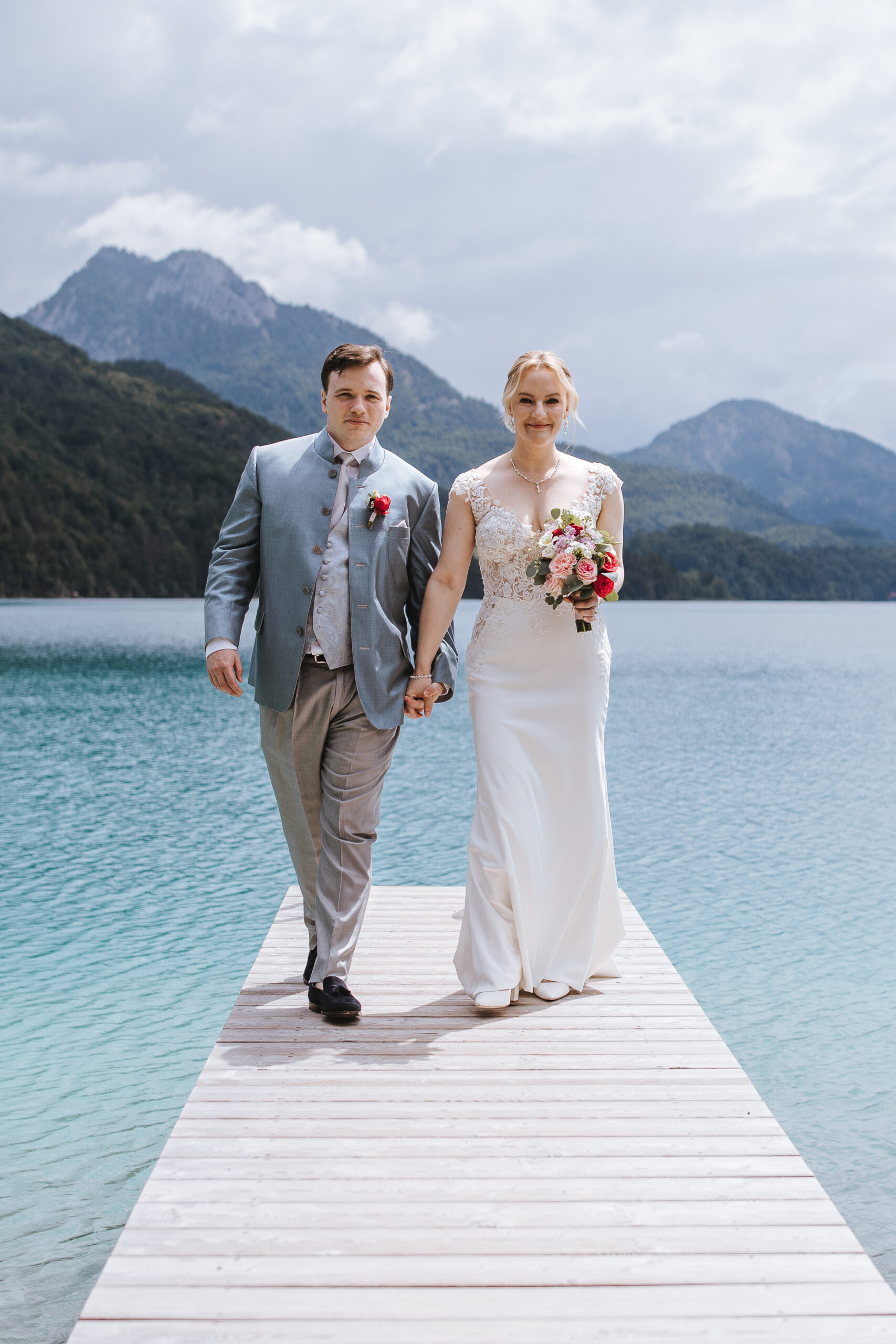 Ein Brautpaar spaziert Hand in Hand auf einem Steg am Seeufer entlang, im Hintergrund ragen Berge empor – eine malerische Hochzeit in Österreich.