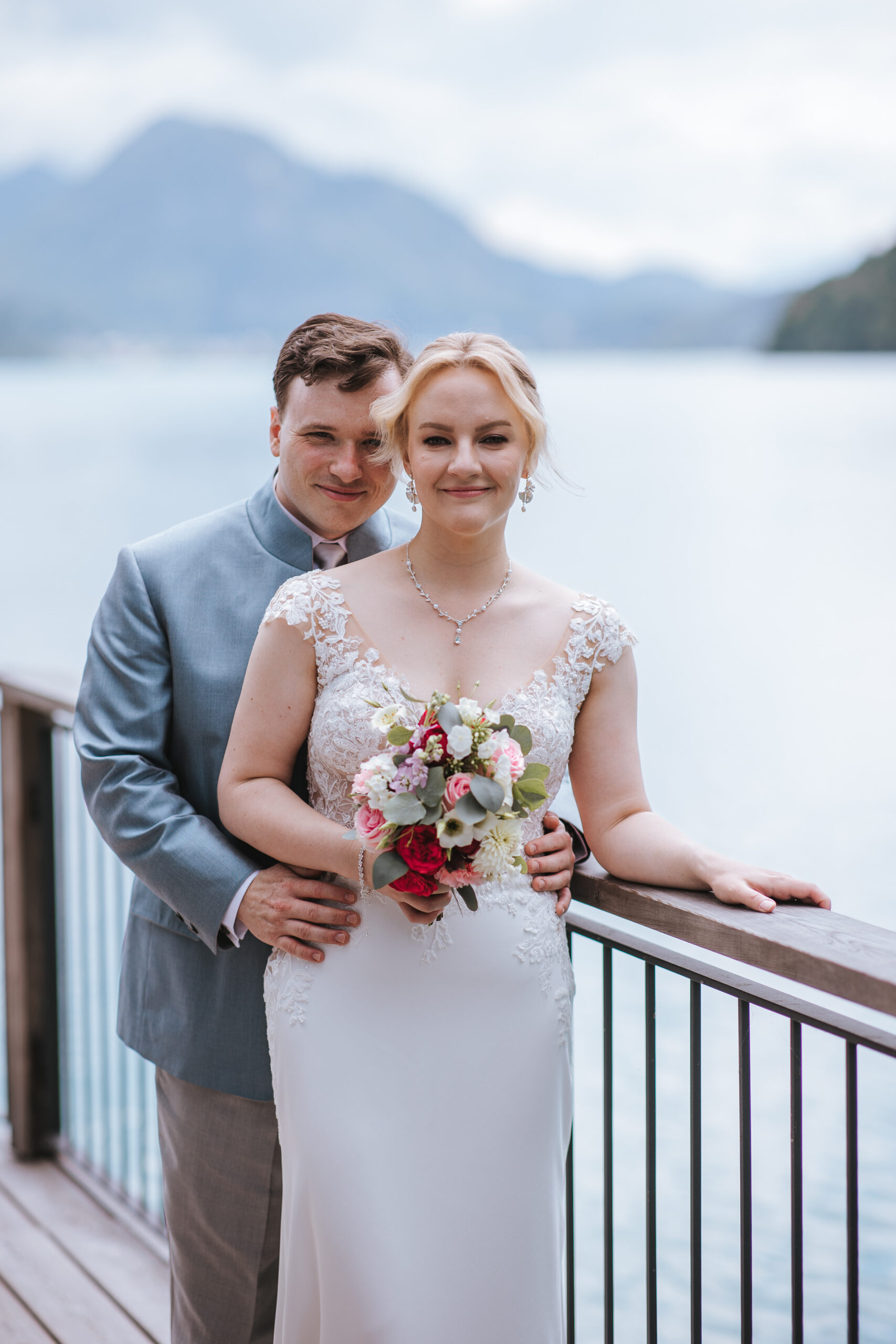 Das lächelnde Brautpaar steht an einem Geländer am Seeufer, im Hintergrund sind Berge zu sehen – perfekt für eine romantische Hochzeit in Österreich.