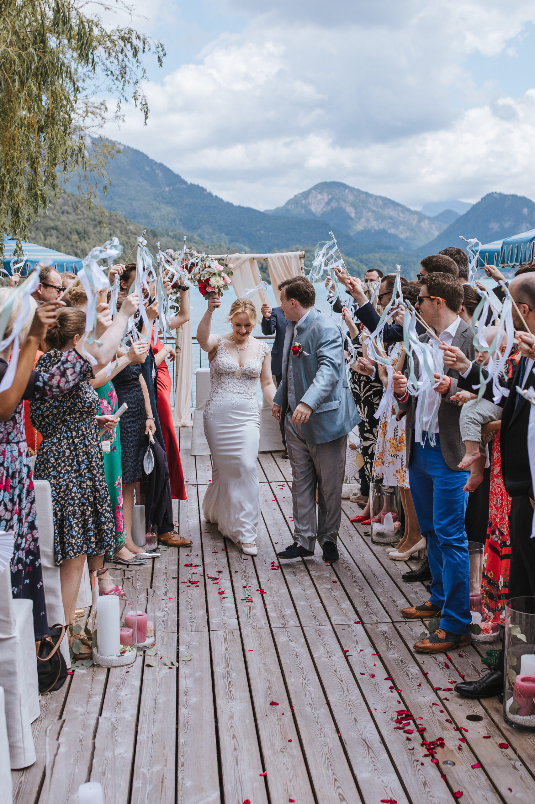 Braut und Bräutigam schreiten in Österreich einen Gang im Freien entlang, während die Gäste unter bewölktem Himmel jubeln und mit Bandstäbchen winken – so entsteht eine zauberhafte Atmosphäre einer Hochzeit im Ausland.