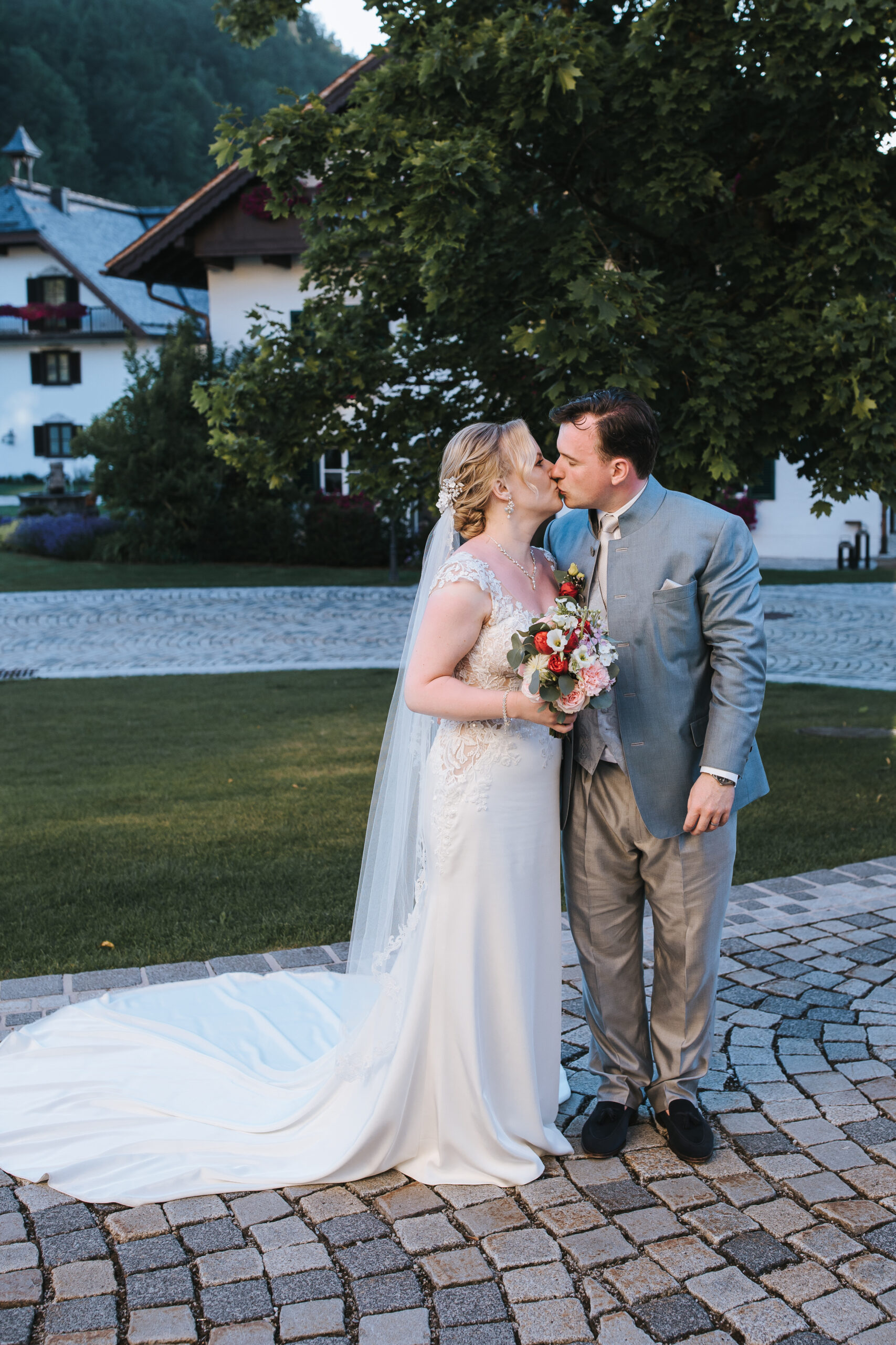 Braut und Bräutigam in Hochzeitskleidung küssen sich im Freien auf einem Steinweg, umgeben von Grün und Gebäuden – ein Bild, das die Romantik einer Hochzeit im Ausland in Österreich einfängt.