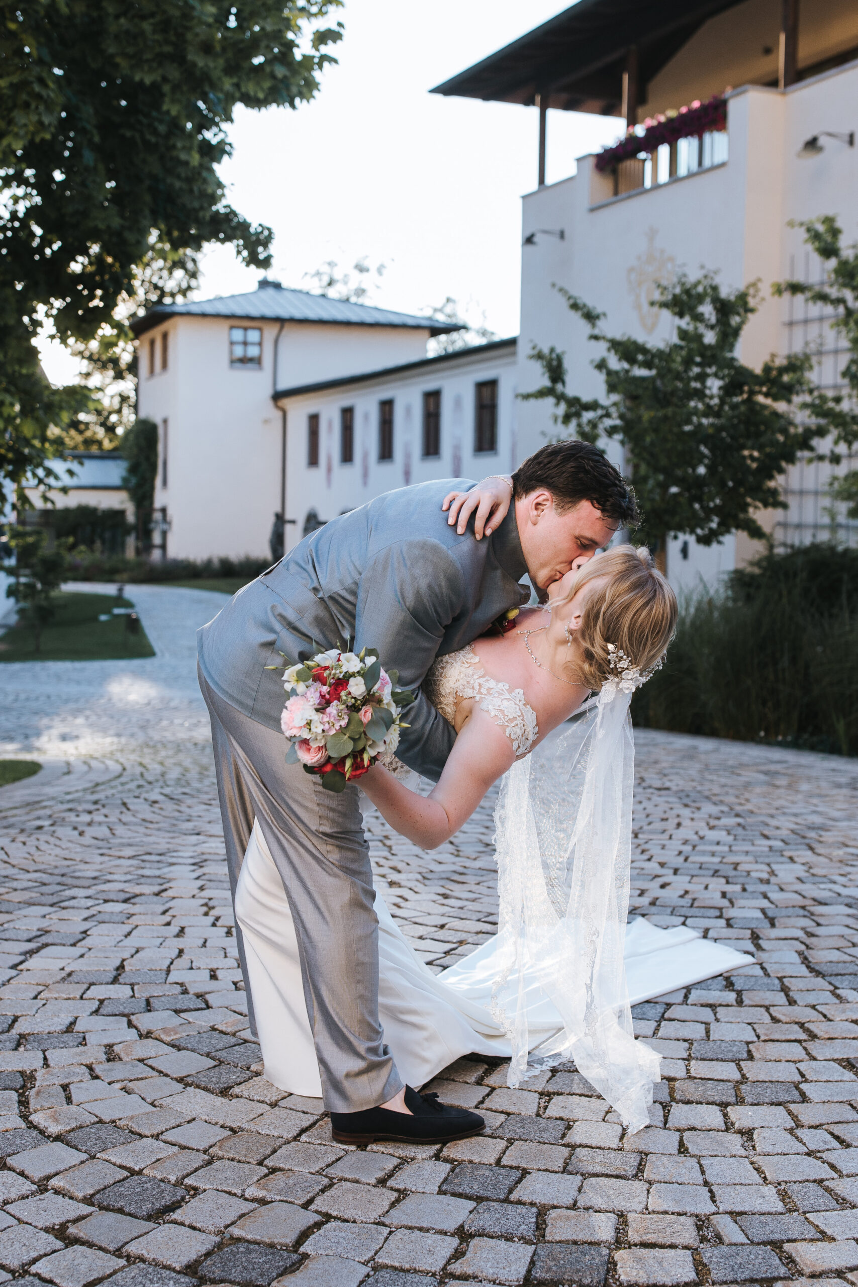 Der Bräutigam im grauen Anzug beugt sich vor und küsst die Braut im weißen Kleid, die einen Blumenstrauß in der Hand hält, auf einem Kopfsteinpflasterweg im Freien bei ihrer romantischen Hochzeit in Österreich.