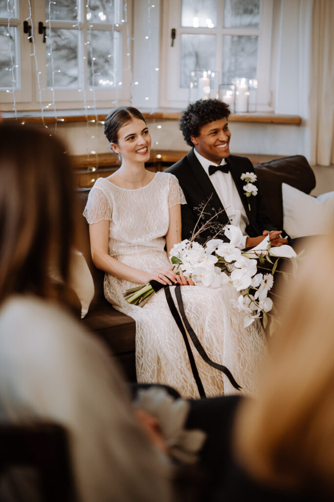 Ein Brautpaar sitzt während der Trauung lächelnd im Innenraum und hält einen weißen Blumenstrauß in den Händen – an einer wunderschönen Hochzeitslocation ohne Catering, die sich perfekt für eine individuelle Hochzeitsplanung eignet.
