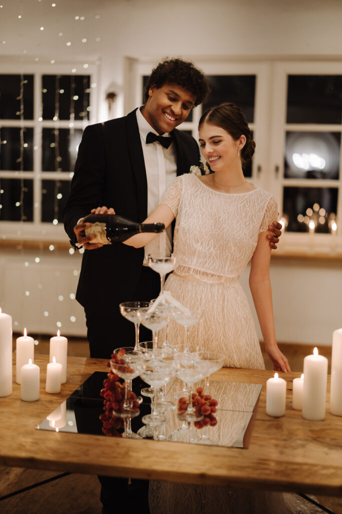 An einer Hochzeitslocation ohne Catering schenkt ein Mann im Smoking einer lächelnden Frau im Abendkleid neben einem Turm aus Gläsern, umgeben von Kerzen, Champagner ein – ideal für Paare, die bei ihrer Hochzeitsplanung auf ein externes Catering setzen.