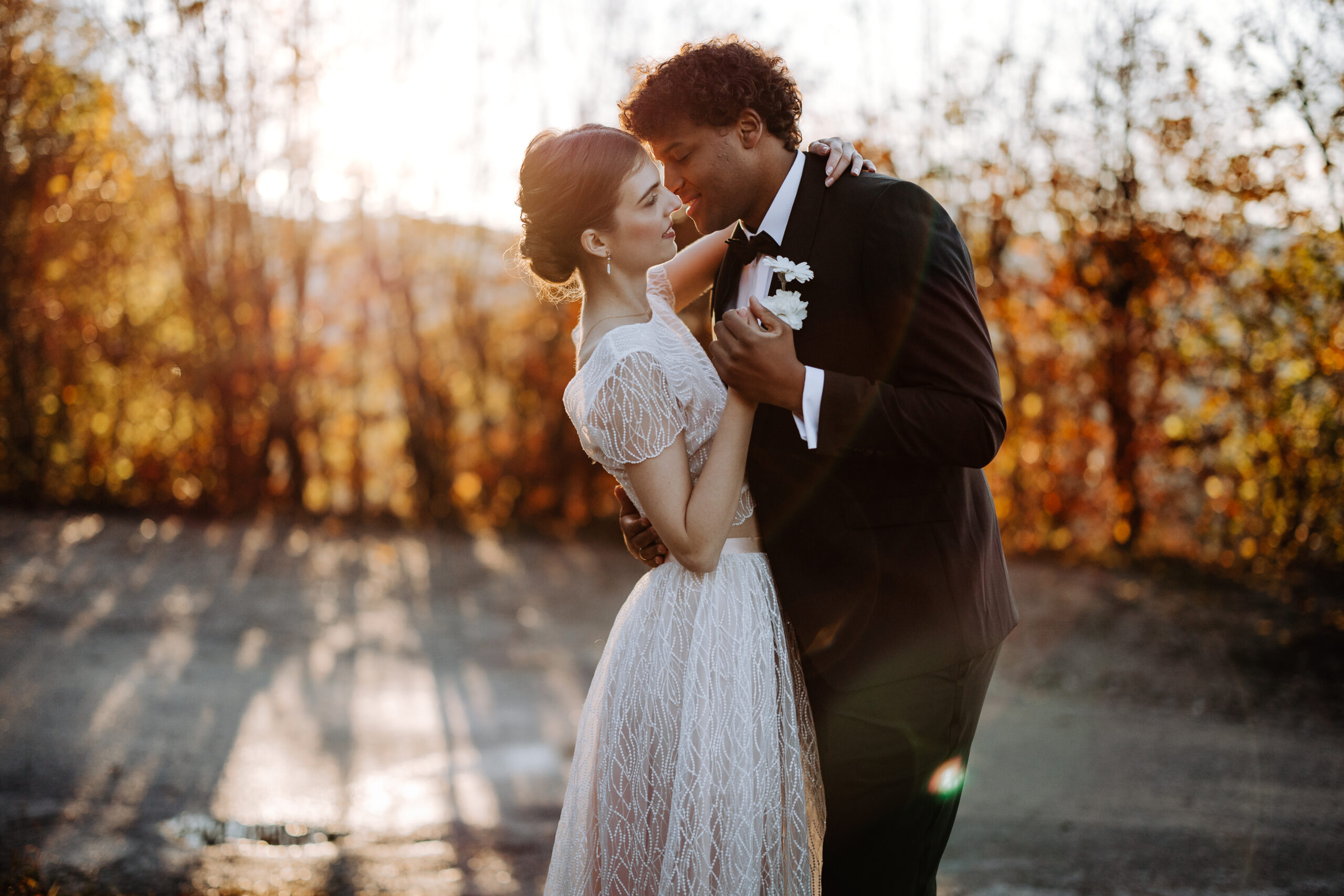 Ein Brautpaar umarmt sich und lächelt sich im Freien bei Sonnenuntergang an, umgeben von herbstlich gefärbten Bäumen – ein idealer Moment für alle, die eine Hochzeitslocation ohne Catering suchen, die eine flexible Hochzeitsplanung und externes Catering ermöglicht.