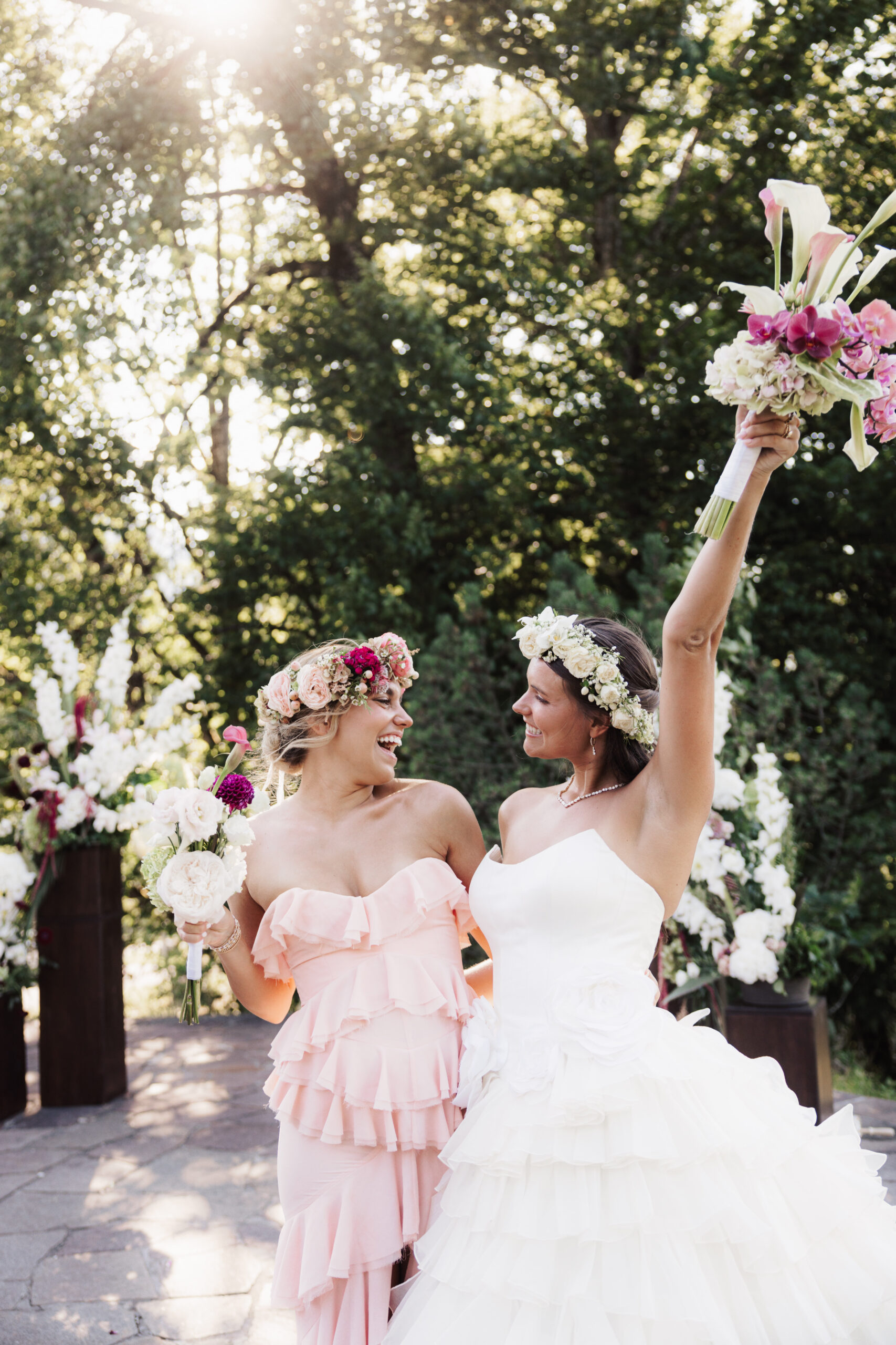 Zwei Frauen in Kleidern und mit Blumenkränzen auf dem Kopf lächeln, halten Blumensträuße in den Händen und feiern draußen im Sonnenschein – und genießen dabei die Vorteile, dass ein Hochzeitsplaner alle wichtigen Aufgaben hinter den Kulissen übernimmt.