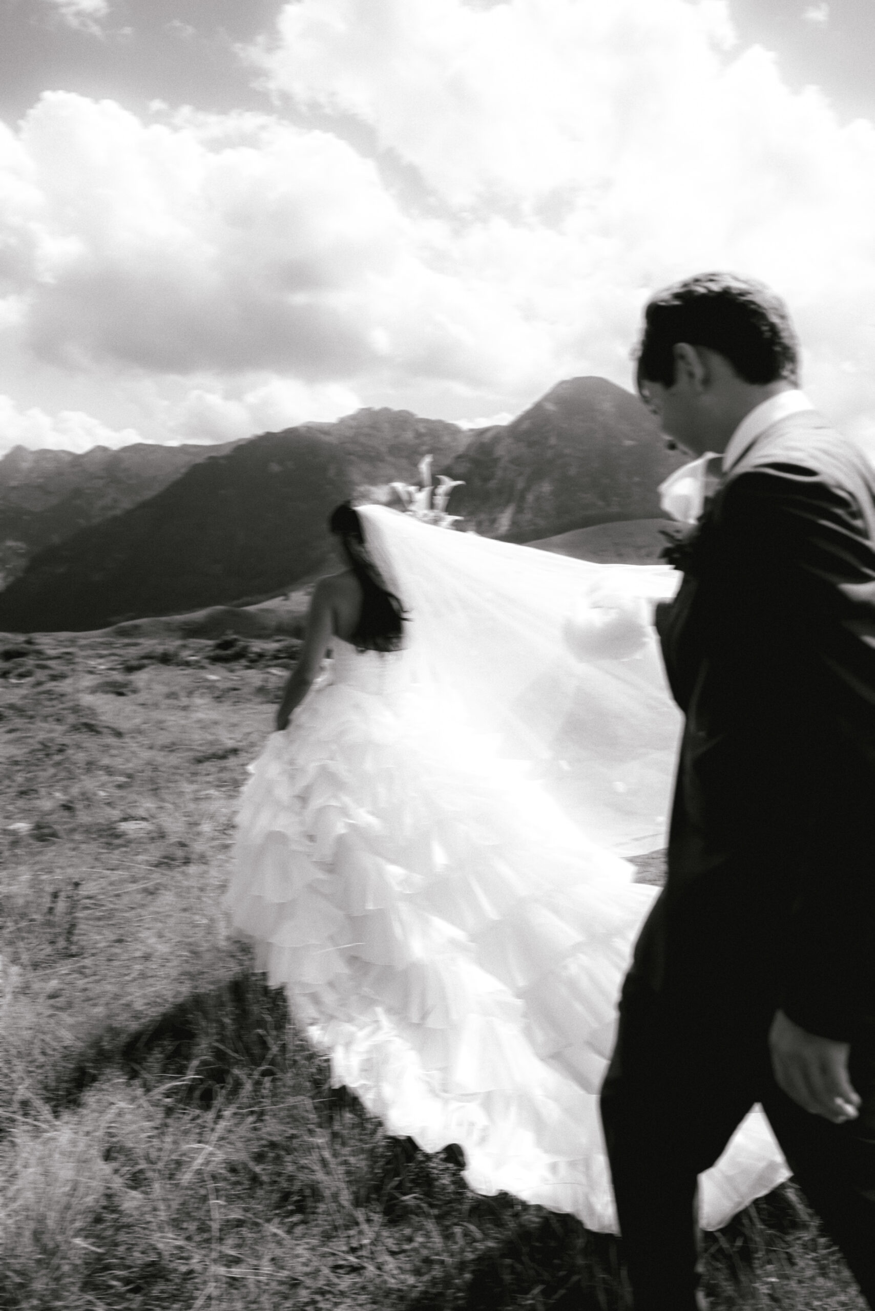 Ein Brautpaar spaziert an seiner Hochzeit draußen in den Bergen, ihr Kleid weht hinter ihr her – in Schwarz-Weiß.