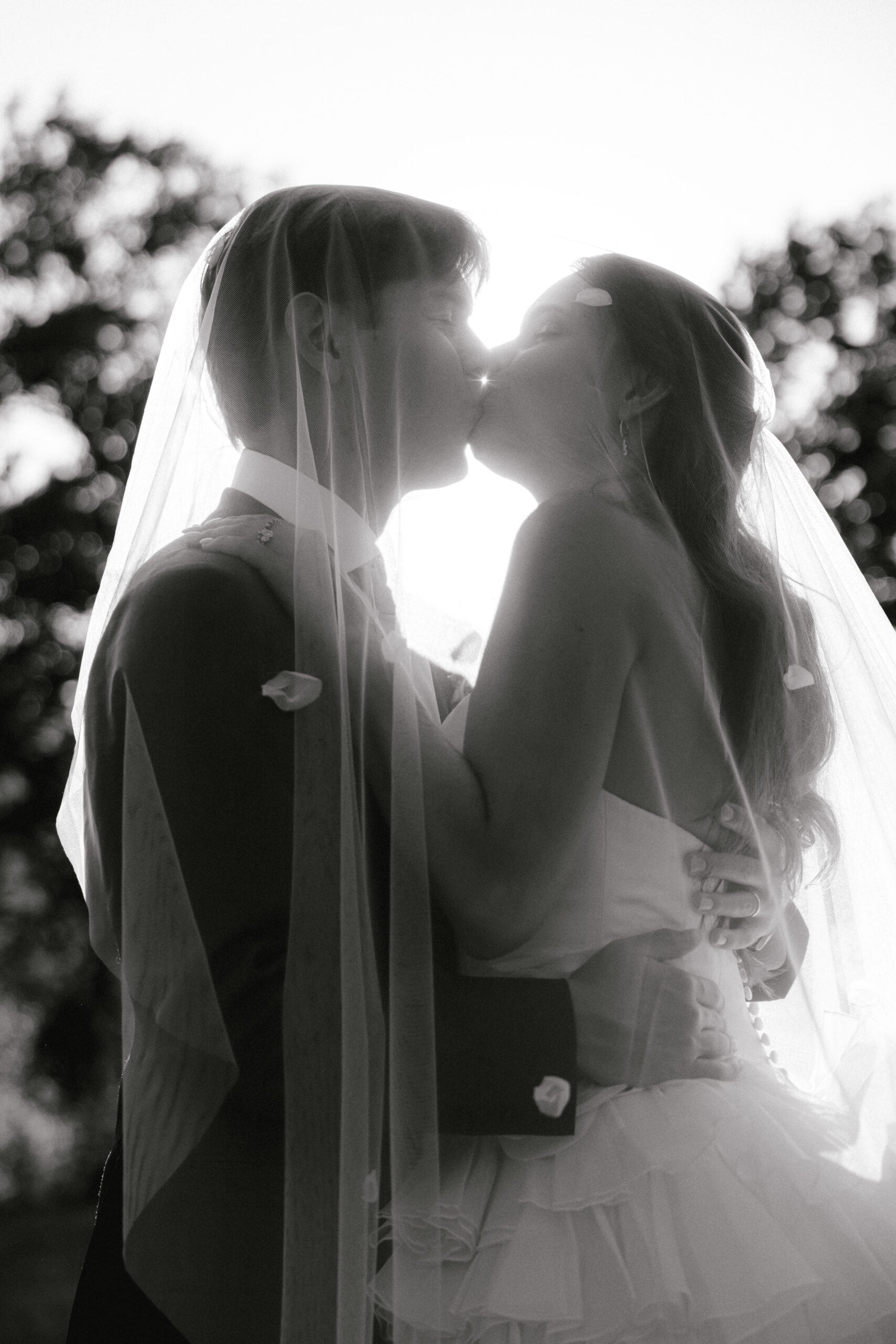 Ein Brautpaar küsst sich im Freien unter einem Schleier, im Gegenlicht der Sonne und umgeben von Blütenblättern – so werden die Magie und Romantik eingefangen, die jede Hochzeit verdient, dank der sorgfältigen Aufgaben, die Ihr Hochzeitsmanager nahtlos koordiniert.