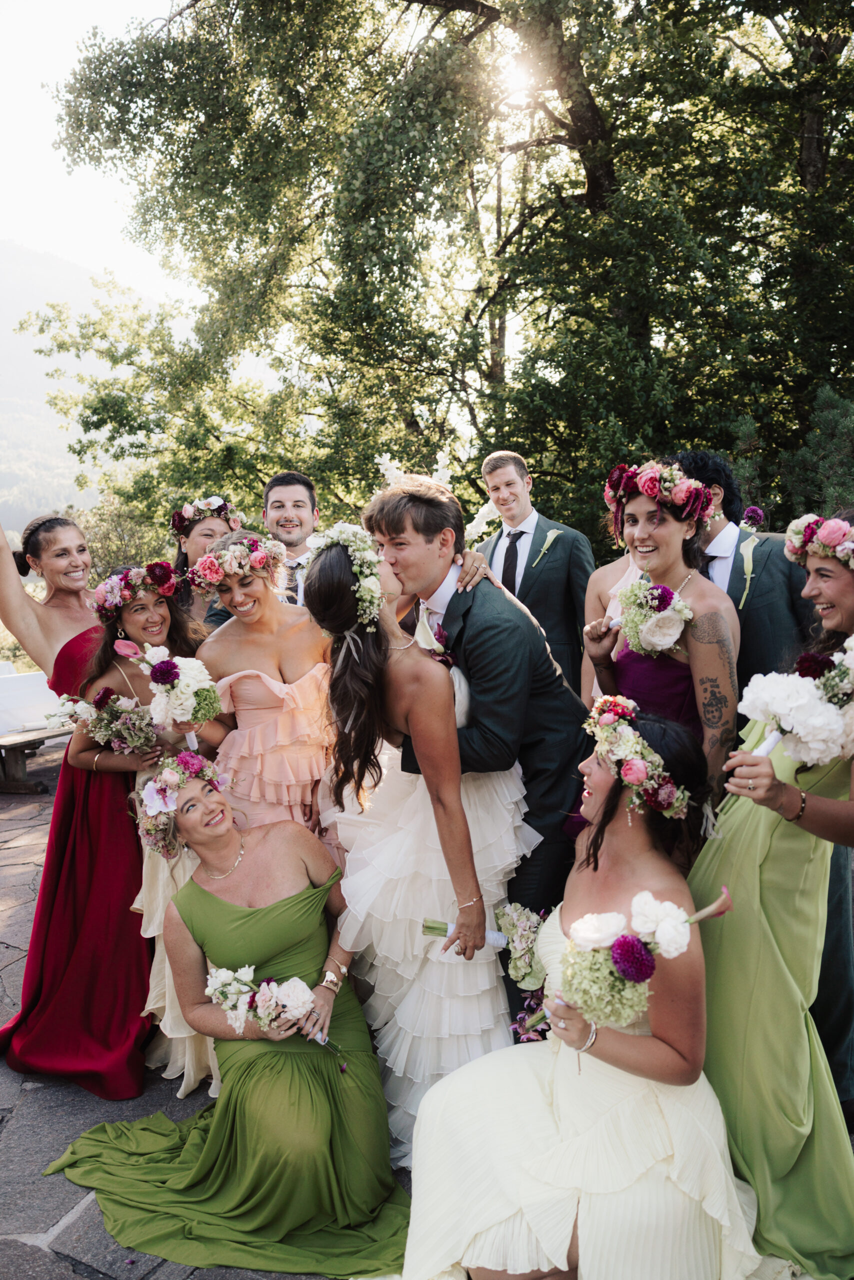 Die Hochzeitsgesellschaft posiert im Freien; Brautjungfern in bunten Kleidern und mit Blumenkränzen, das Brautpaar küsst sich, strahlende Gesichter – alles perfekt aufeinander abgestimmt dank der Aufgaben und Vorteile eines Wedding Day Managers.