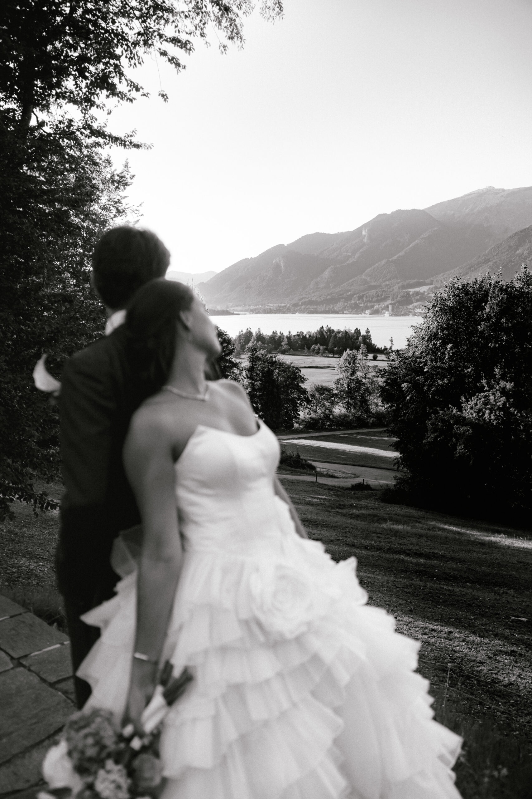 Das Brautpaar in Hochzeitskleidung blickt auf eine malerische Berg- und Seelandschaft, fotografiert in Schwarz-Weiß – ein zeitloser Moment, der von ihrem Hochzeitskoordinator perfekt inszeniert wurde, der dafür sorgte, dass jedes Detail der Hochzeit makellos war.