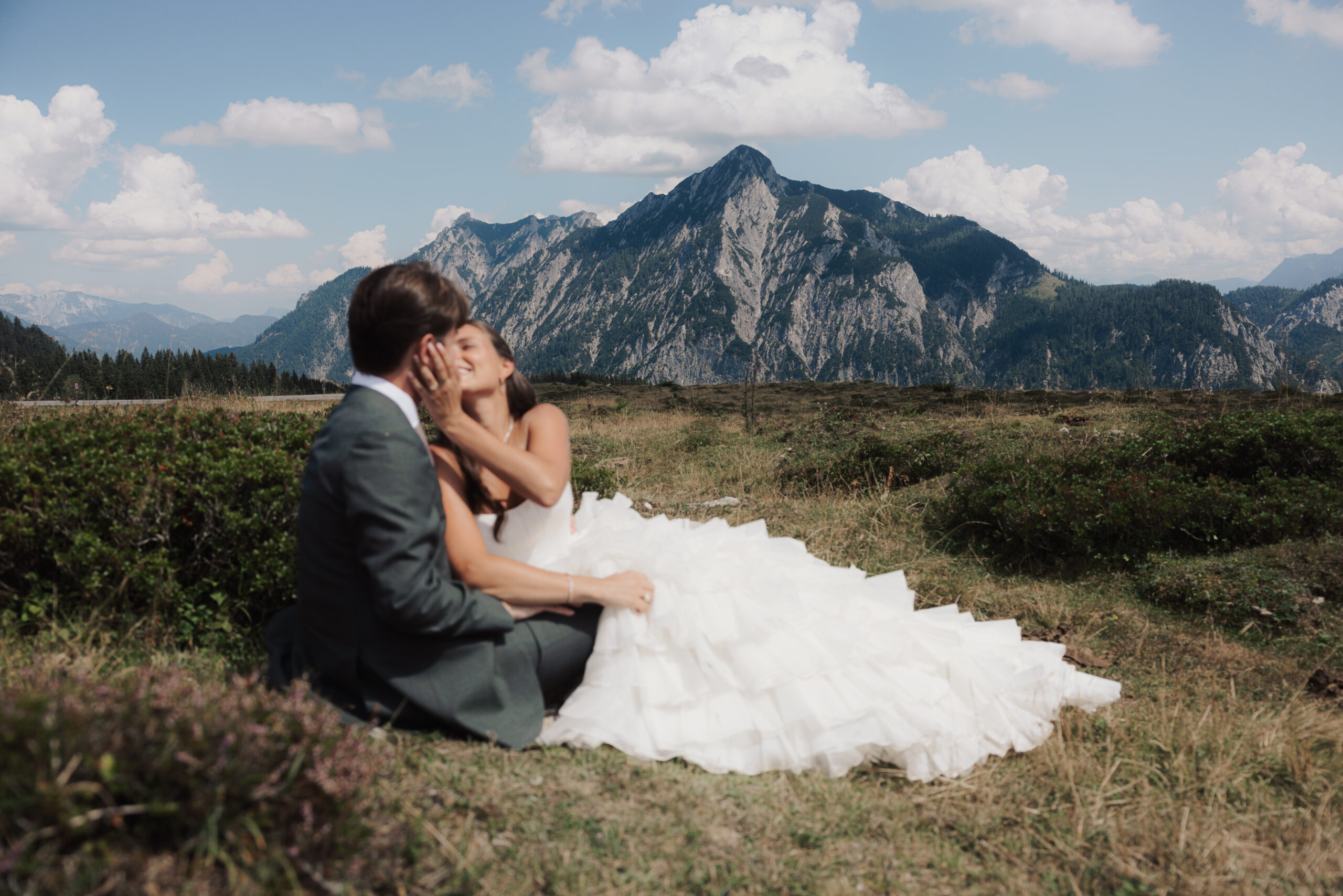 Braut und Bräutigam sitzen sich im Gras in den Armen liegend gegenüber, im Hintergrund sind Berge und der blaue Himmel zu sehen – ein perfekter Moment, festgehalten an ihrer Hochzeit, die von ihrem Wedding Day Manager sorgfältig koordiniert wurde, um sicherzustellen, dass alle Aufgaben reibungslos erledigt wurden.
