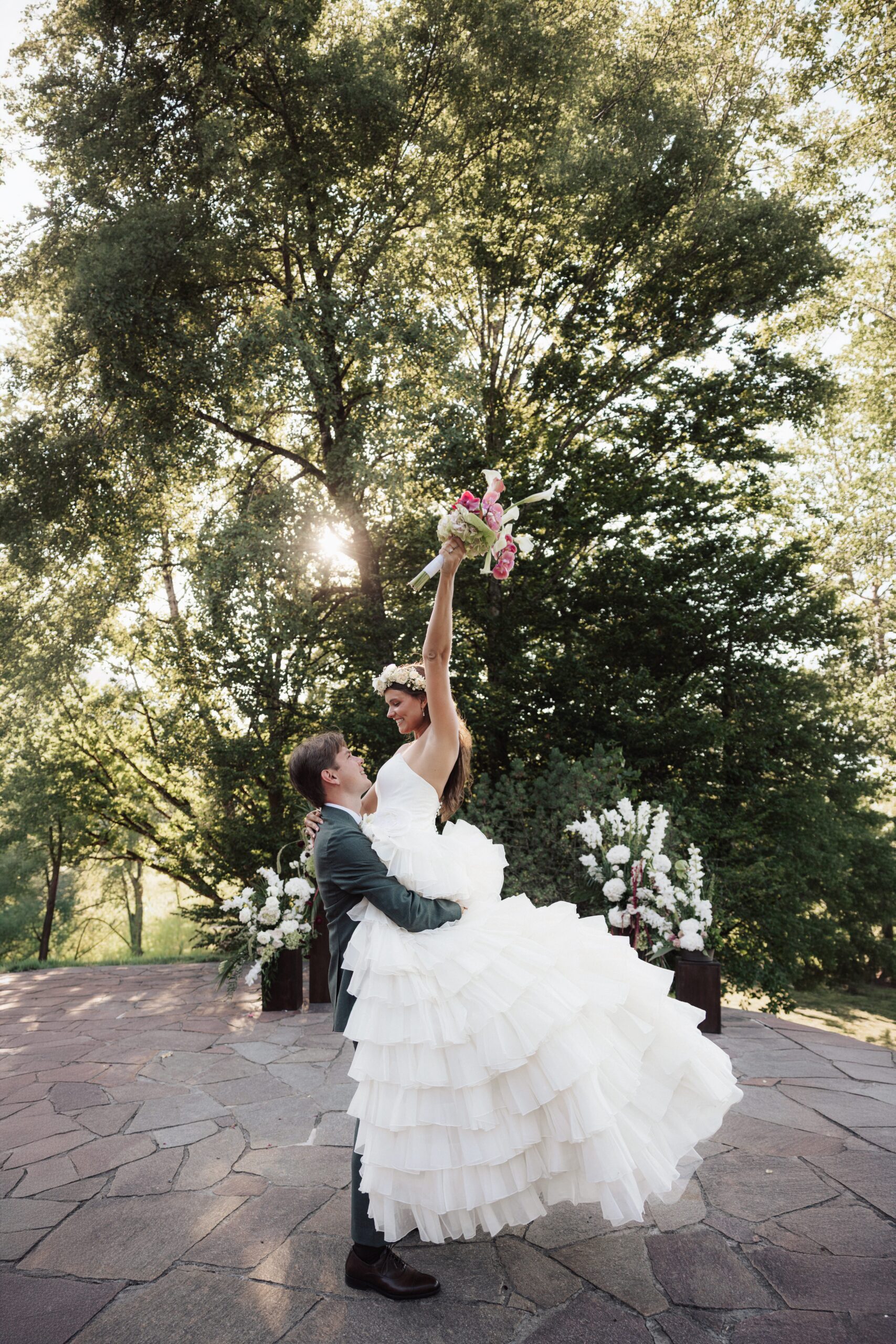 Ein Bräutigam hebt seine Braut im weißen Kleid hoch, während sie einen Blumenstrauß in der Hand hält – umgeben von Bäumen und Blumen im Freien: Ein Bild, das die Magie ihrer Hochzeit perfekt einfängt.