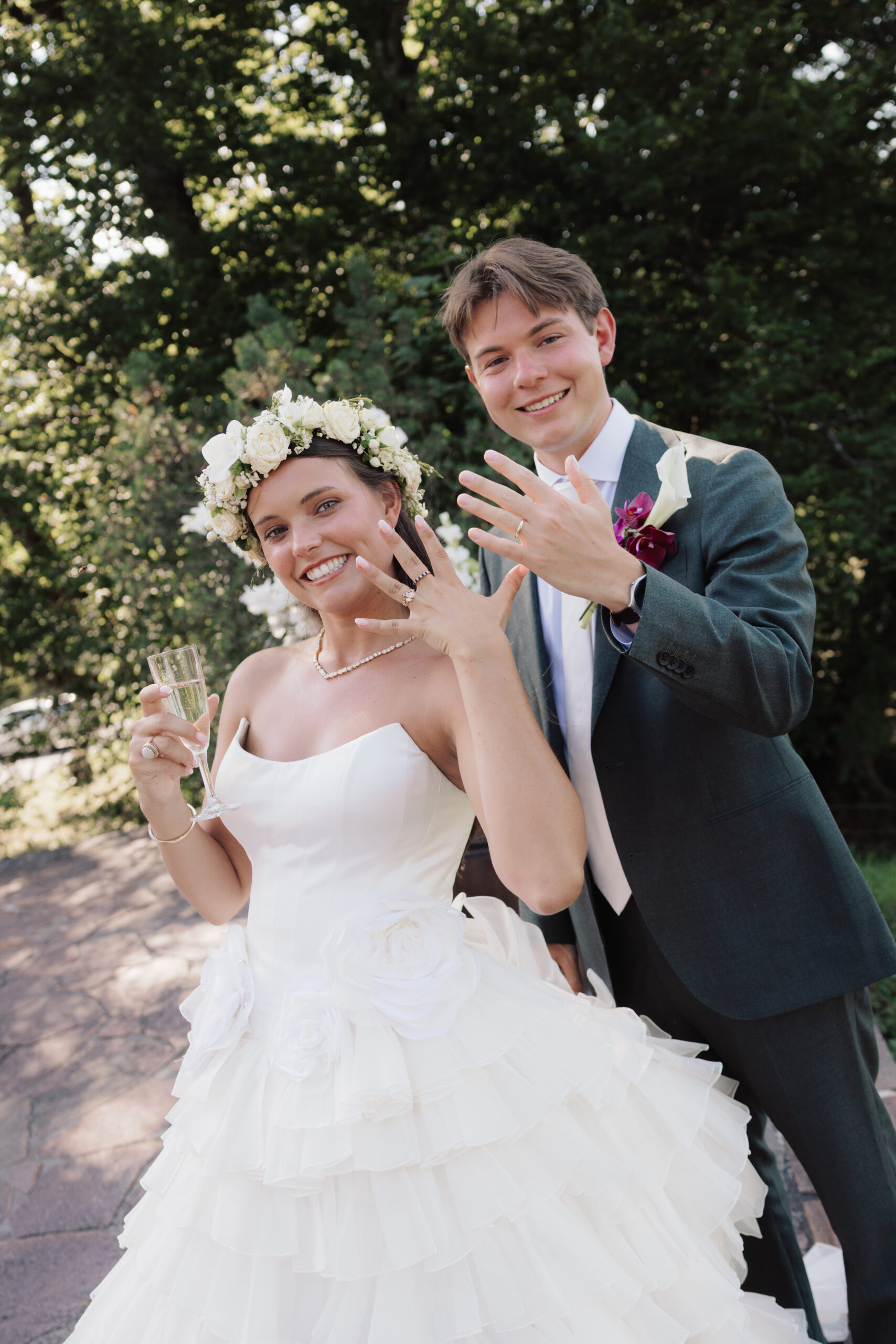 Das lächelnde Brautpaar zeigt seine Eheringe, die Braut hält ein Glas Champagner in der Hand, im Hintergrund ist eine grüne Landschaft zu sehen – ein fröhlicher Hochzeitsmoment, der durch den Wedding Day Manager nahtlos in Szene gesetzt wurde, der dafür sorgte, dass alle Aufgaben perfekt koordiniert wurden.