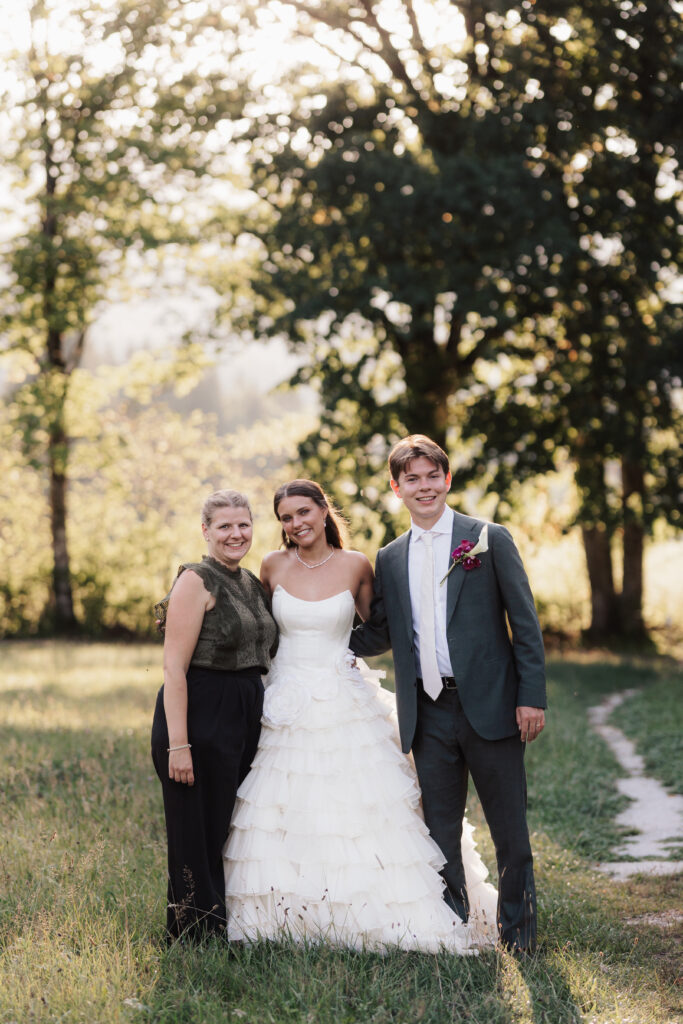 Drei Personen stehen gemeinsam im Freien: eine lächelnde Braut in einem weißen Kleid, ein Bräutigam und eine Frau in dunkler Kleidung, die wie eine engagierte Hochzeitskoordinatorin alles im Blick hat und dafür sorgt, dass alle Aufgaben für diese wunderschöne Hochzeit perfekt erledigt werden.