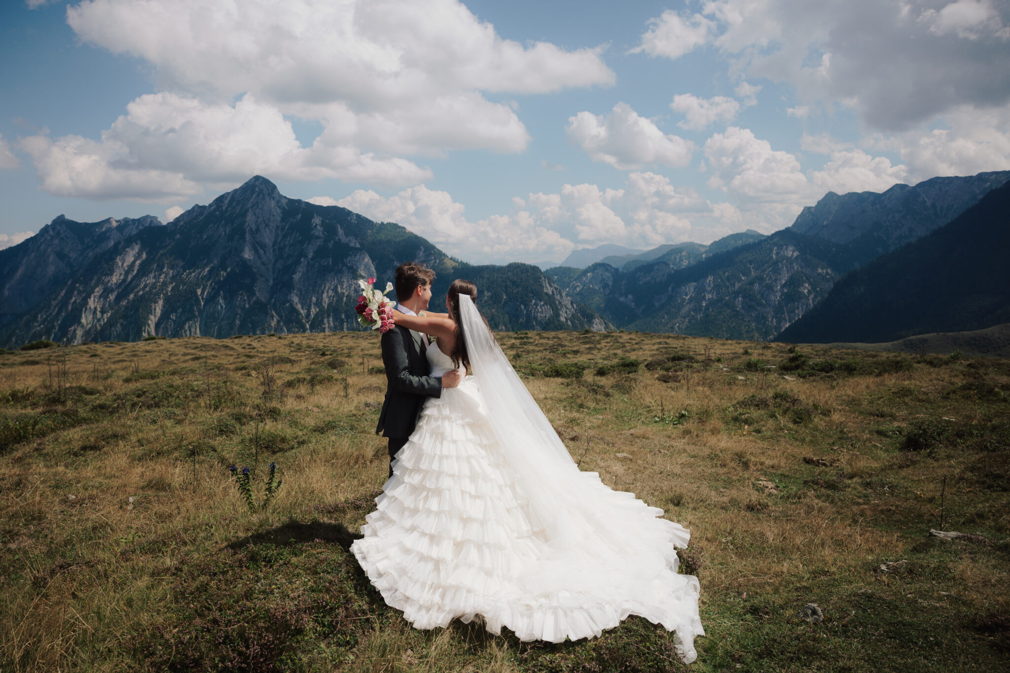 Braut und Bräutigam umarmen sich im Freien vor dem Hintergrund der Berge unter einem teilweise bewölkten Himmel, während ihr Hochzeitskoordinator dafür sorgt, dass alle Hochzeitsaufgaben reibungslos erledigt werden. Stressfreie Hochzeit in Österreich