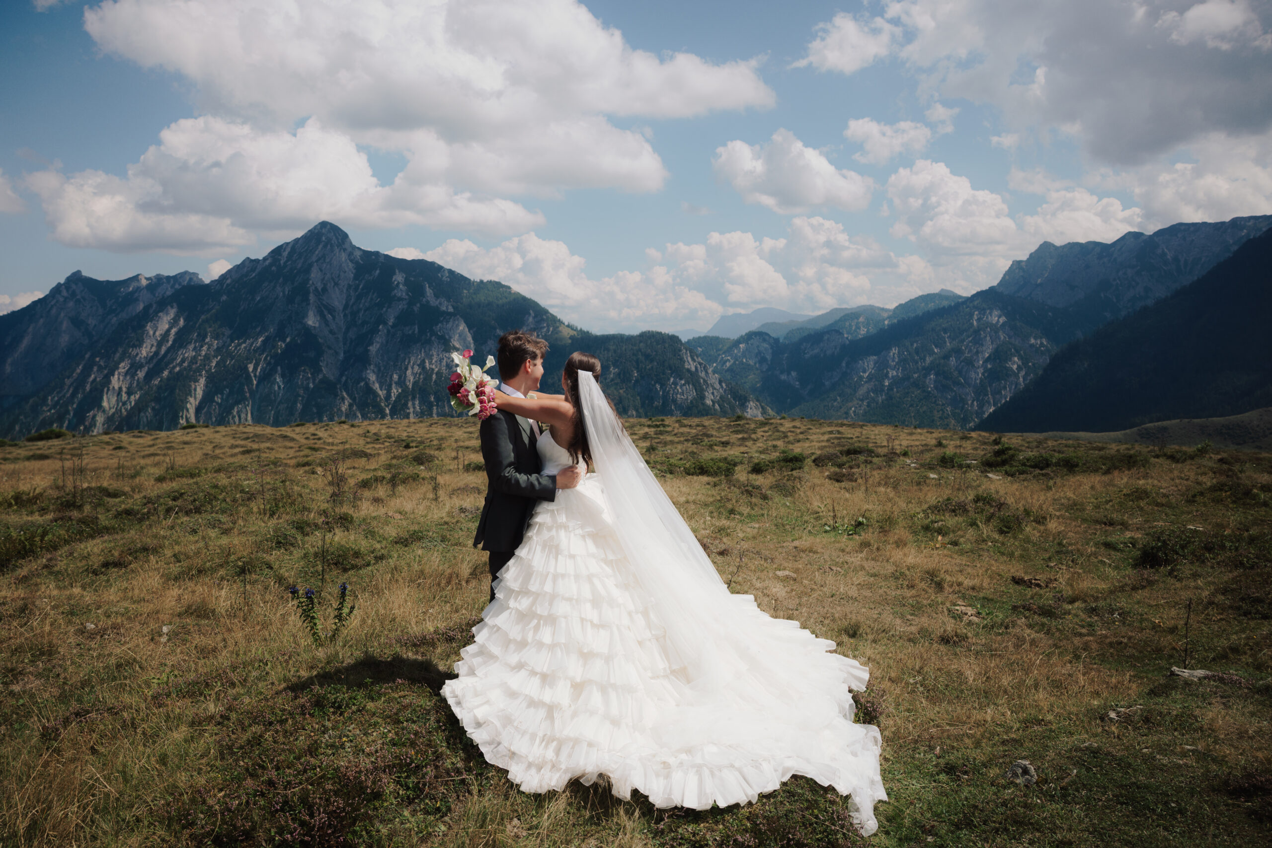Braut und Bräutigam umarmen sich im Freien vor dem Hintergrund der Berge unter einem teilweise bewölkten Himmel, während ihr Hochzeitskoordinator dafür sorgt, dass alle Hochzeitsaufgaben reibungslos erledigt werden. Stressfreie Hochzeit in Österreich
